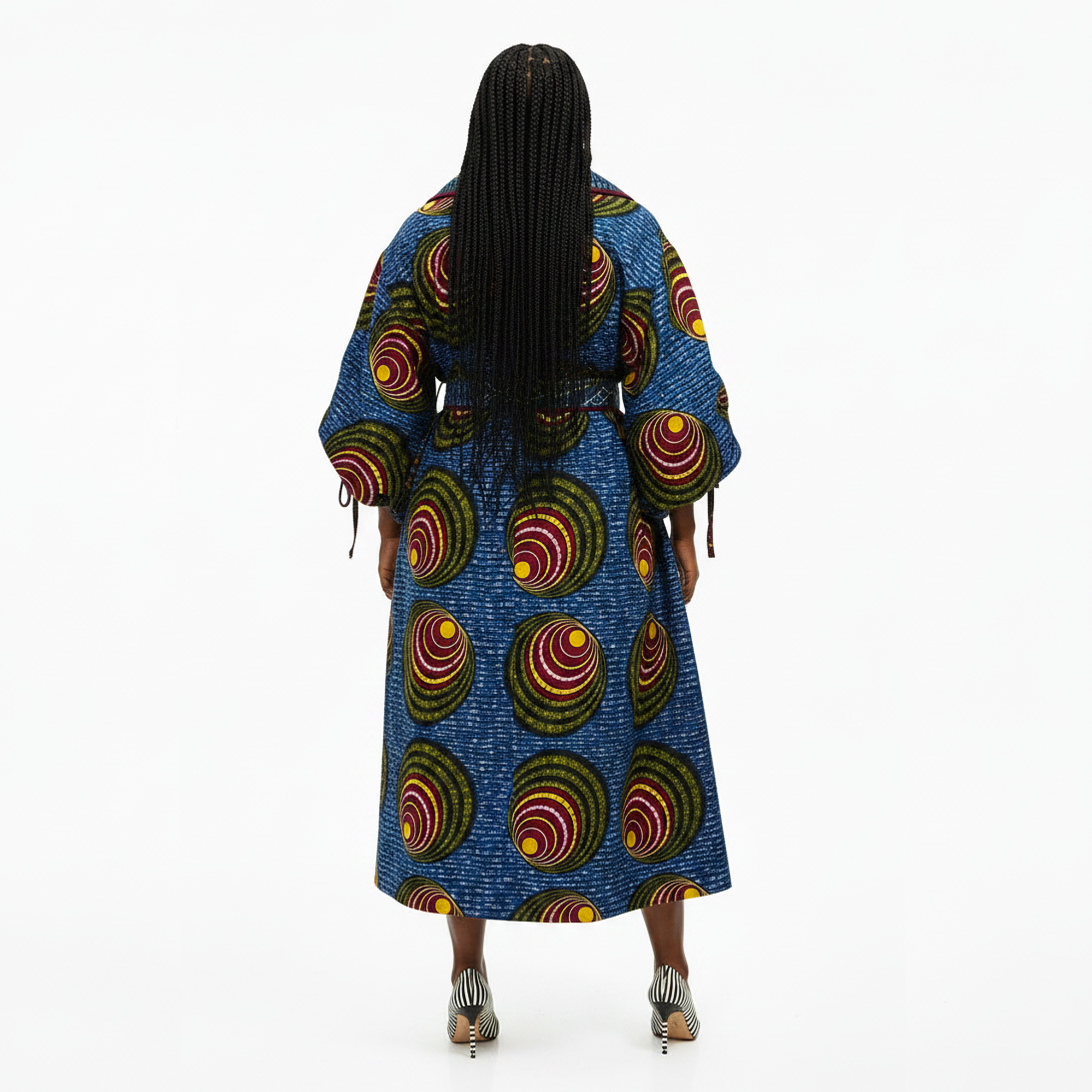 A woman viewed from behind wearing a long blue dress with a bold circular pattern in red, yellow, green, and orange, paired with metallic high heels against a plain white background.