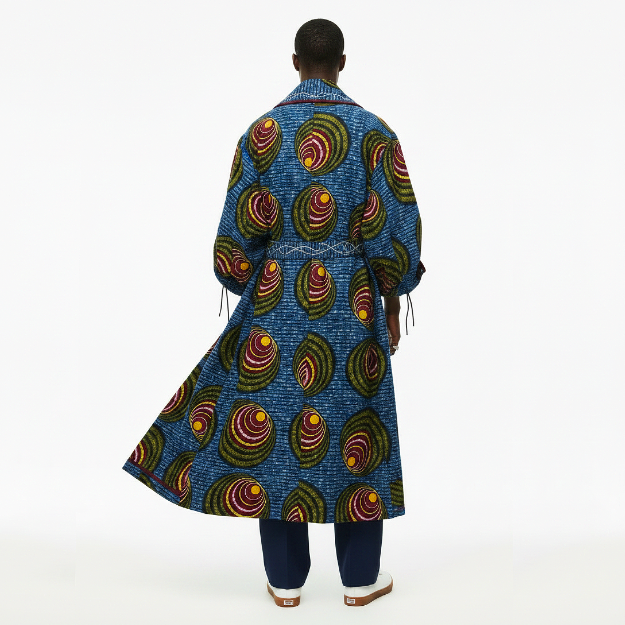 A model viewed from the back wearing a long blue wrap coat with a vibrant multicolor African wax print pattern of concentric circles, paired with navy pants and white shoes, against a plain white background.