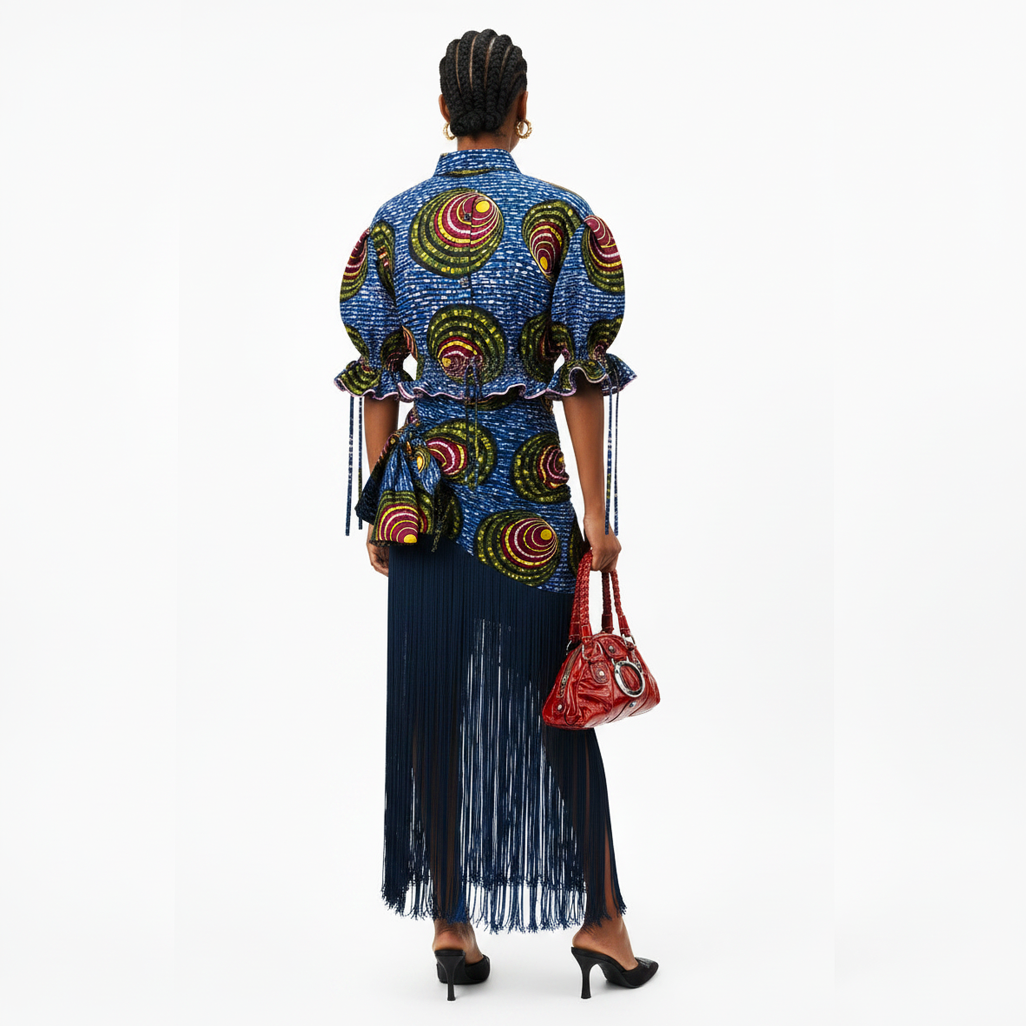 A model is shown from the back wearing a vibrant blue African wax print shirt with colorful circular patterns and a matching fringed skirt. She is holding a red patterned handbag against a plain white studio background.