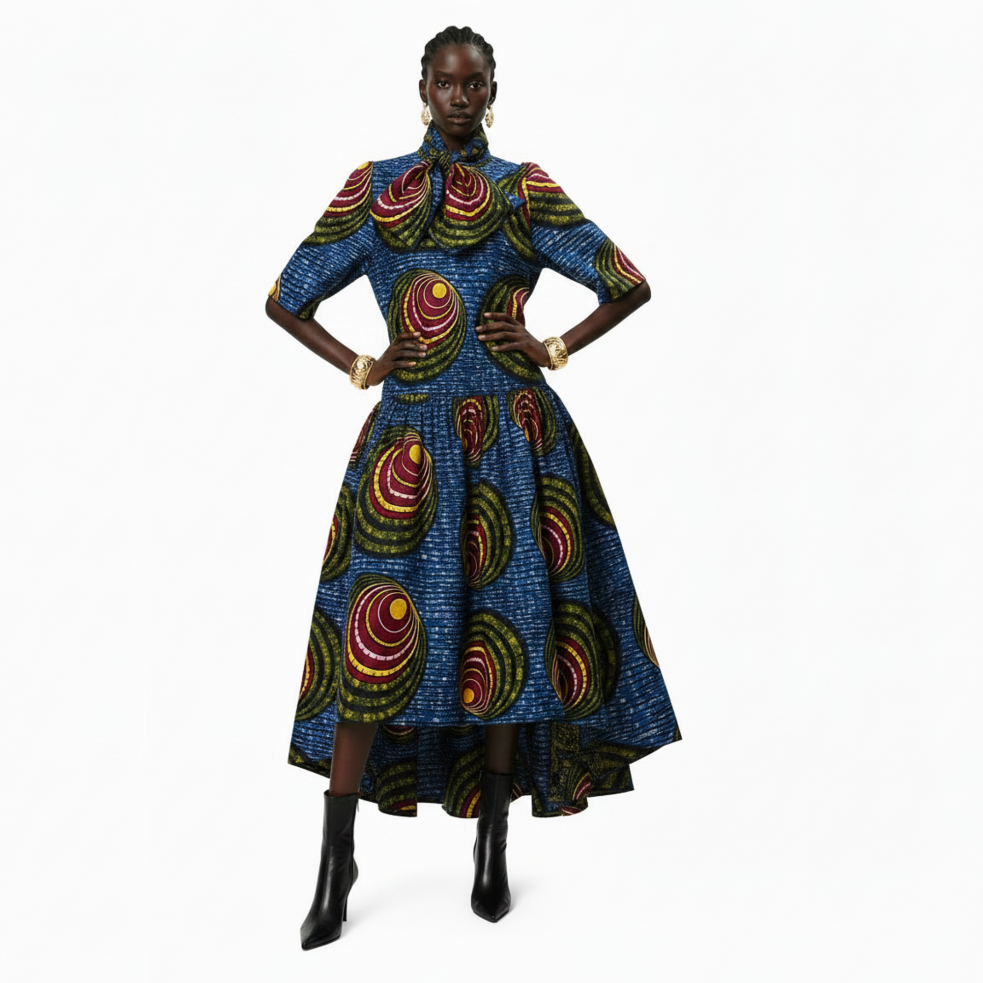 A model wearing a high-low midi dress with a vibrant blue African wax print pattern featuring red, yellow, and green circular motifs, styled with black ankle boots against a plain white background.