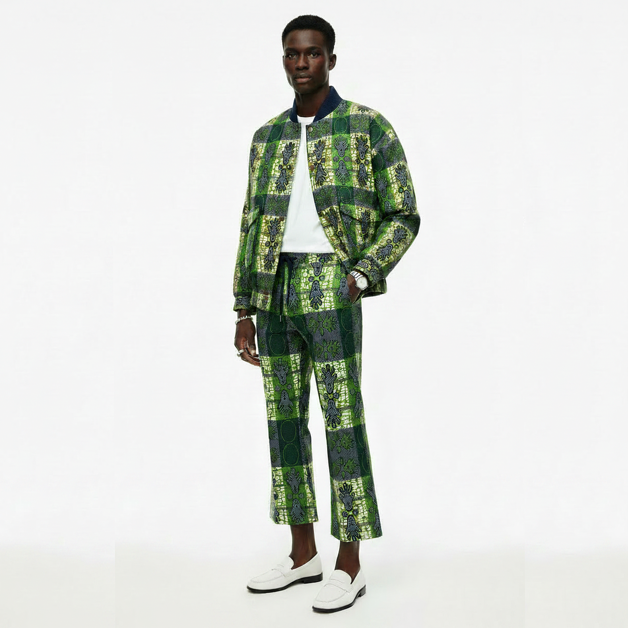 A male model stands against a plain white studio background wearing a matching green and black patterned jacket and trousers set, a white T-shirt, and white loafers.