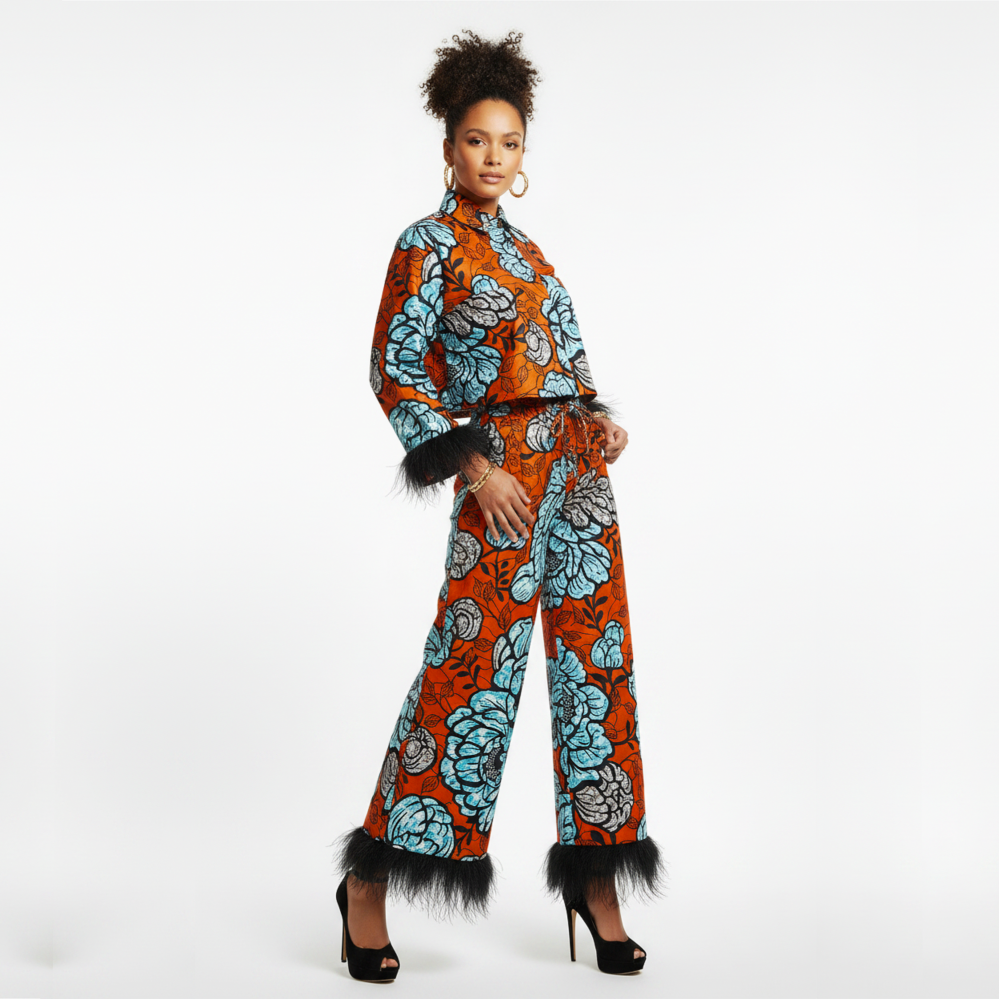 A model wearing a matching two-piece jumpsuit set in vibrant orange and light blue floral African wax print, featuring black feather trim on the cuffs and hems, styled with black high heels against a white studio background.