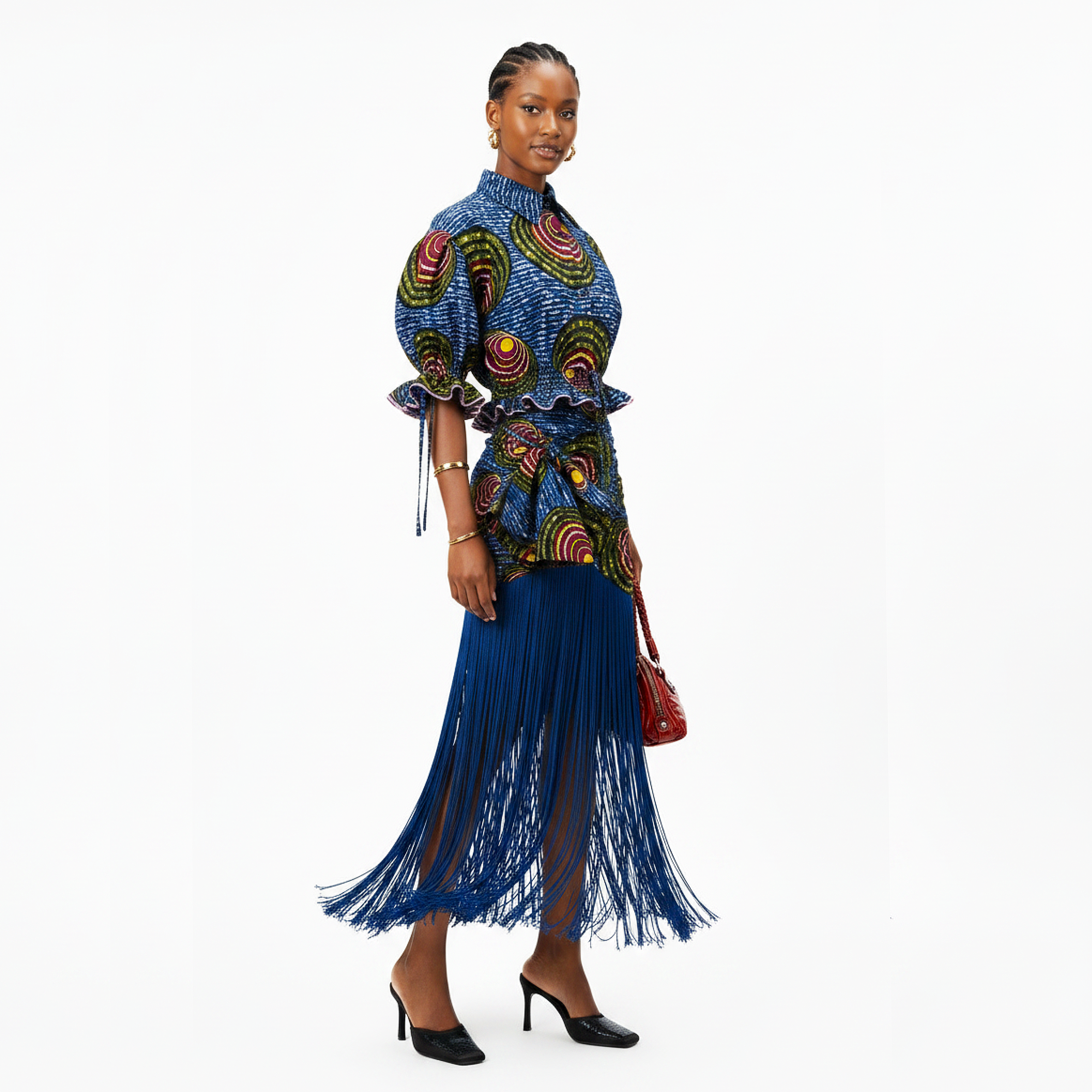 A model is shown from the waist up wearing a vibrant blue and multicolor patterned top with a matching blue fringe skirt, holding a small red handbag against a plain white background.
