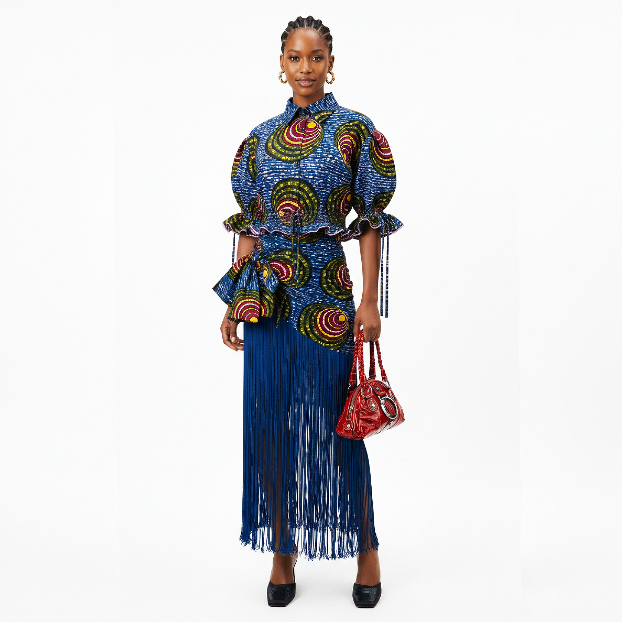 A model stands against a white studio background wearing a blue patterned top and matching long fringe skirt, holding a red handbag.