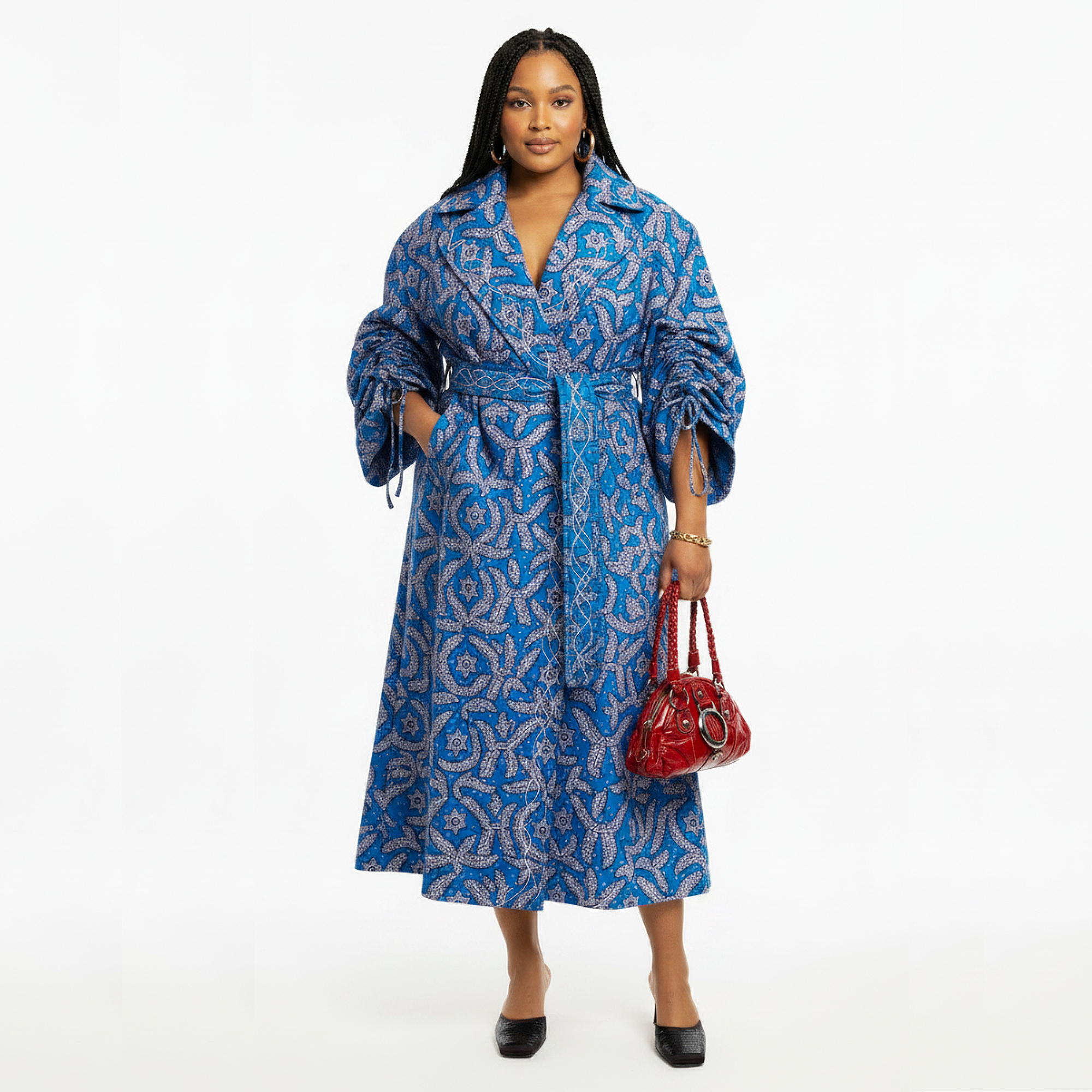 A woman modeling a blue and white patterned midi wrap coat with a tie waist, holding a small red handbag against a plain white studio background.