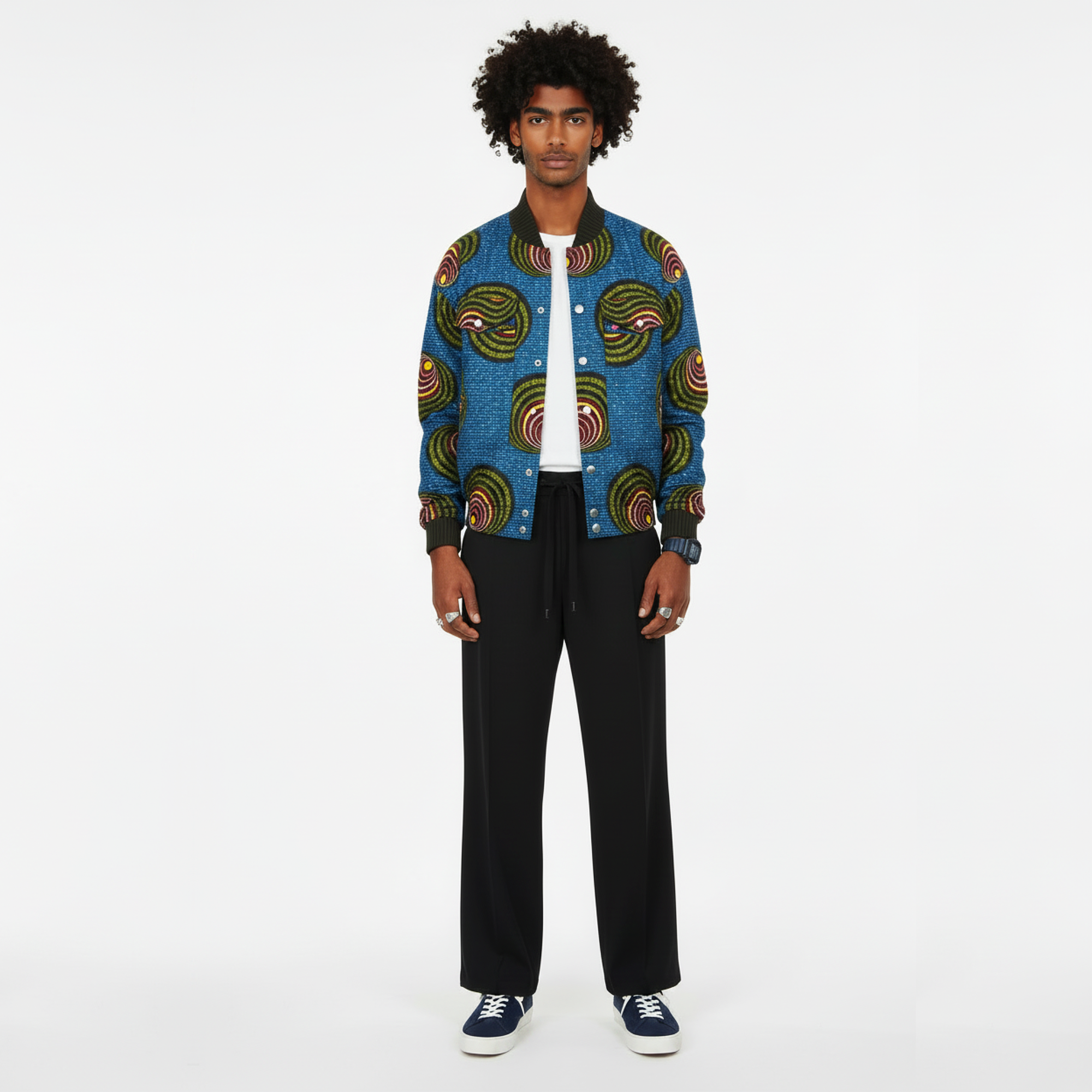 A man stands against a plain white studio background wearing a blue patterned bomber jacket with circular green, yellow, and red motifs, black wide-leg pants, a white T-shirt, and navy sneakers.