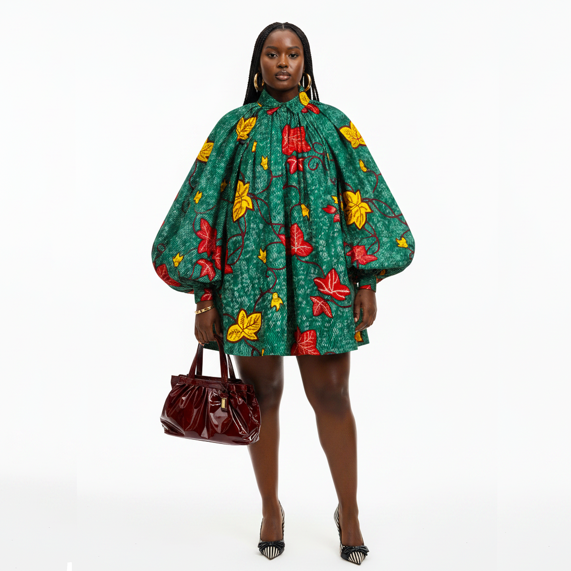 A model stands against a white studio background wearing a green short dress with a red and yellow floral print, voluminous balloon sleeves, and holding a dark red patent leather handbag.