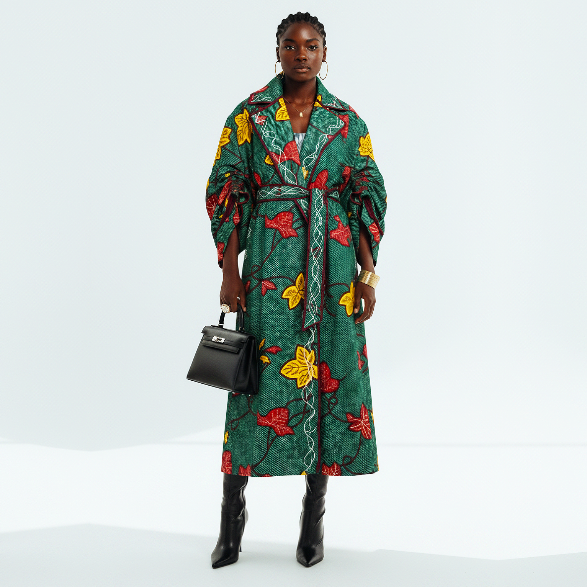 A model stands against a light gray studio background wearing a long green wrap-style coat with a bold red and yellow floral print, holding a small black handbag and wearing black pointed boots.