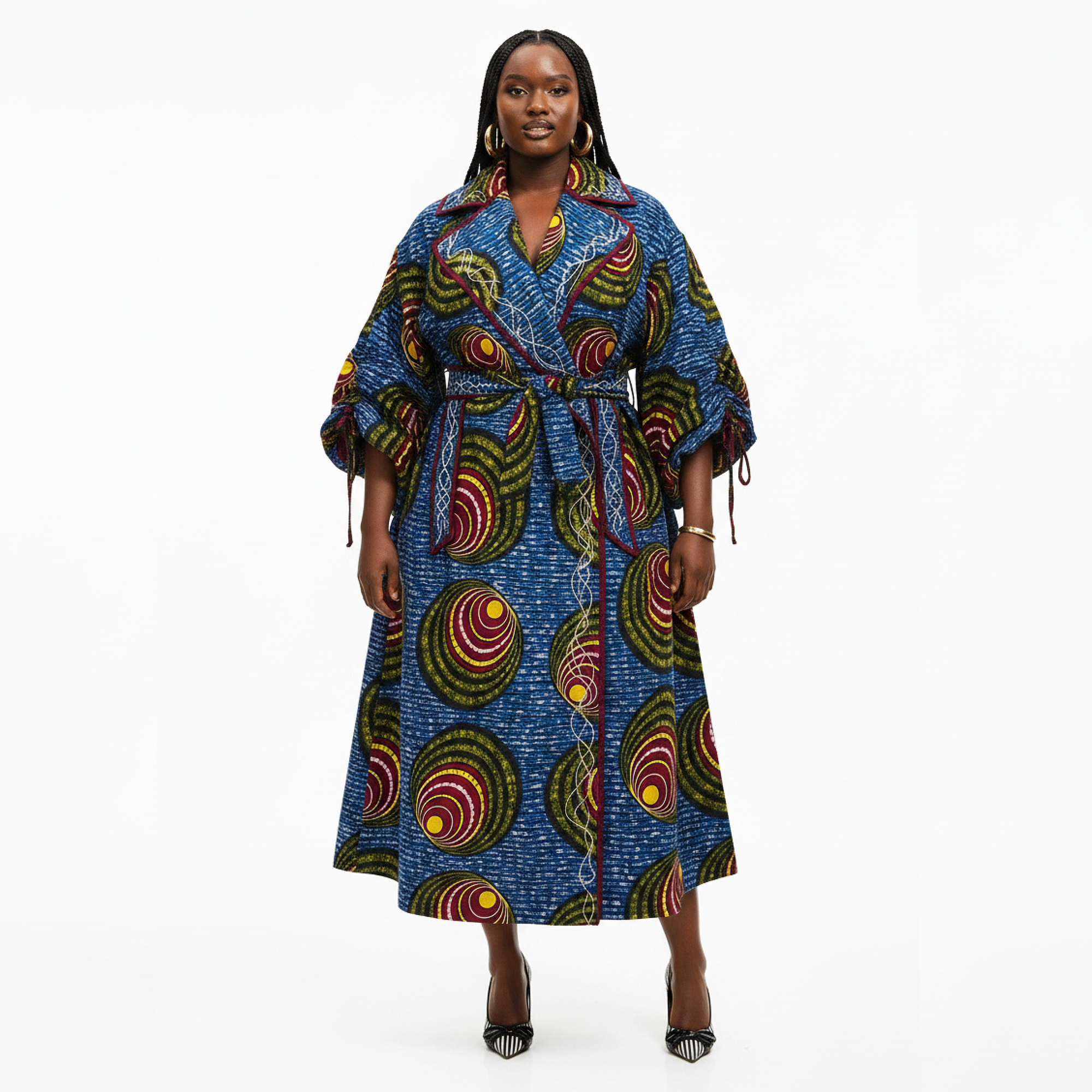 A woman modeling a long blue African wax print wrap dress with a waist tie and multicolor circular patterns, set against a plain white studio background.