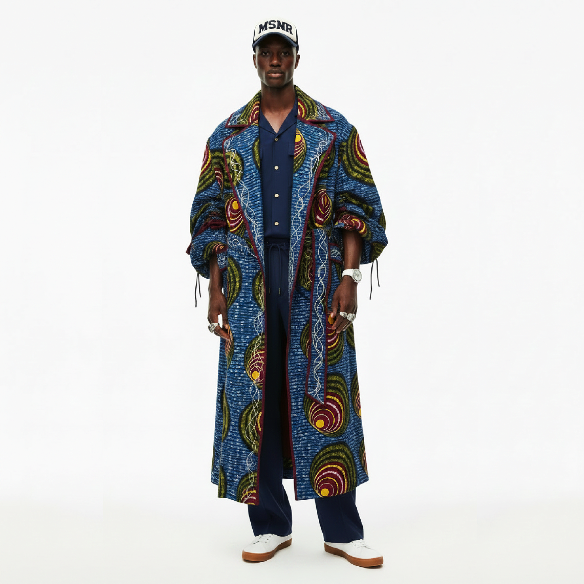 A man modeling a long blue patterned coat with colorful circular motifs, worn over a navy shirt and trousers, paired with a white cap and white sneakers, against a plain white background.