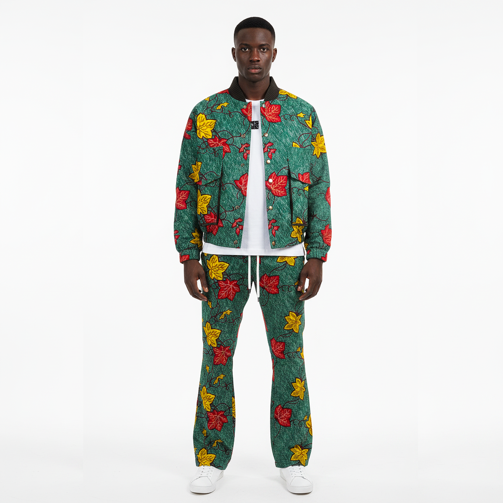 A male model stands against a plain white studio background wearing a matching two-piece tracksuit. The set features a green base with a bold, colorful abstract pattern in red, yellow, and black, styled over a white t-shirt and white sneakers.