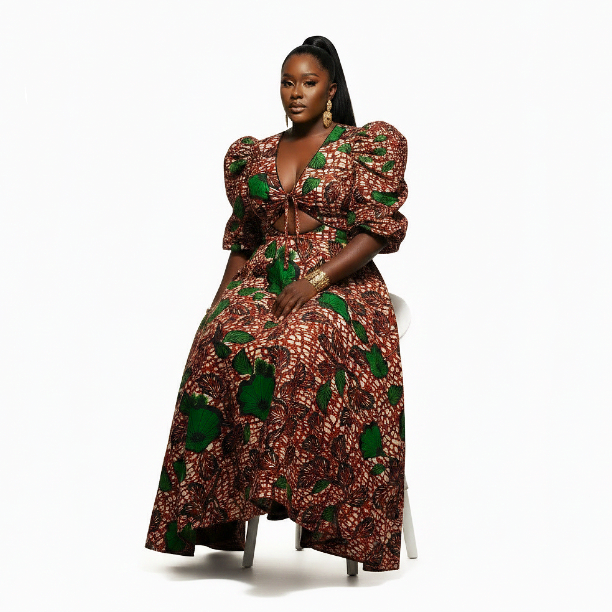 A woman seated on a white chair wearing a voluminous African print dress with puff sleeves, a deep V-neckline, and a brown, red, and green patterned fabric.