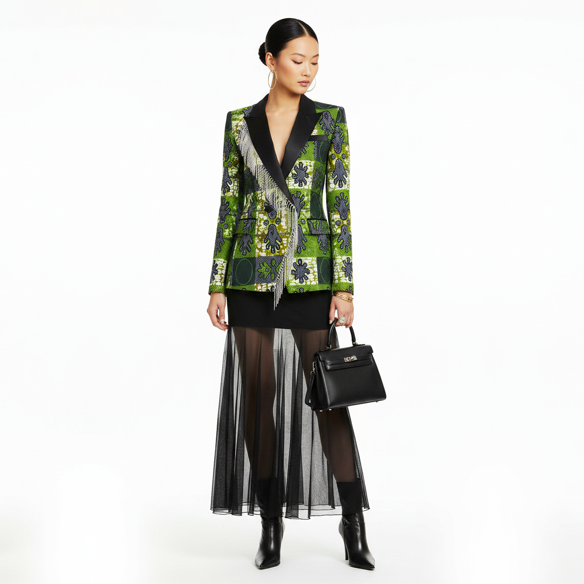 A model stands against a white studio background wearing a green and black patterned blazer with a silver fringe detail, a black mesh midi skirt, sheer black tights, black ankle boots, and holding a black handbag.