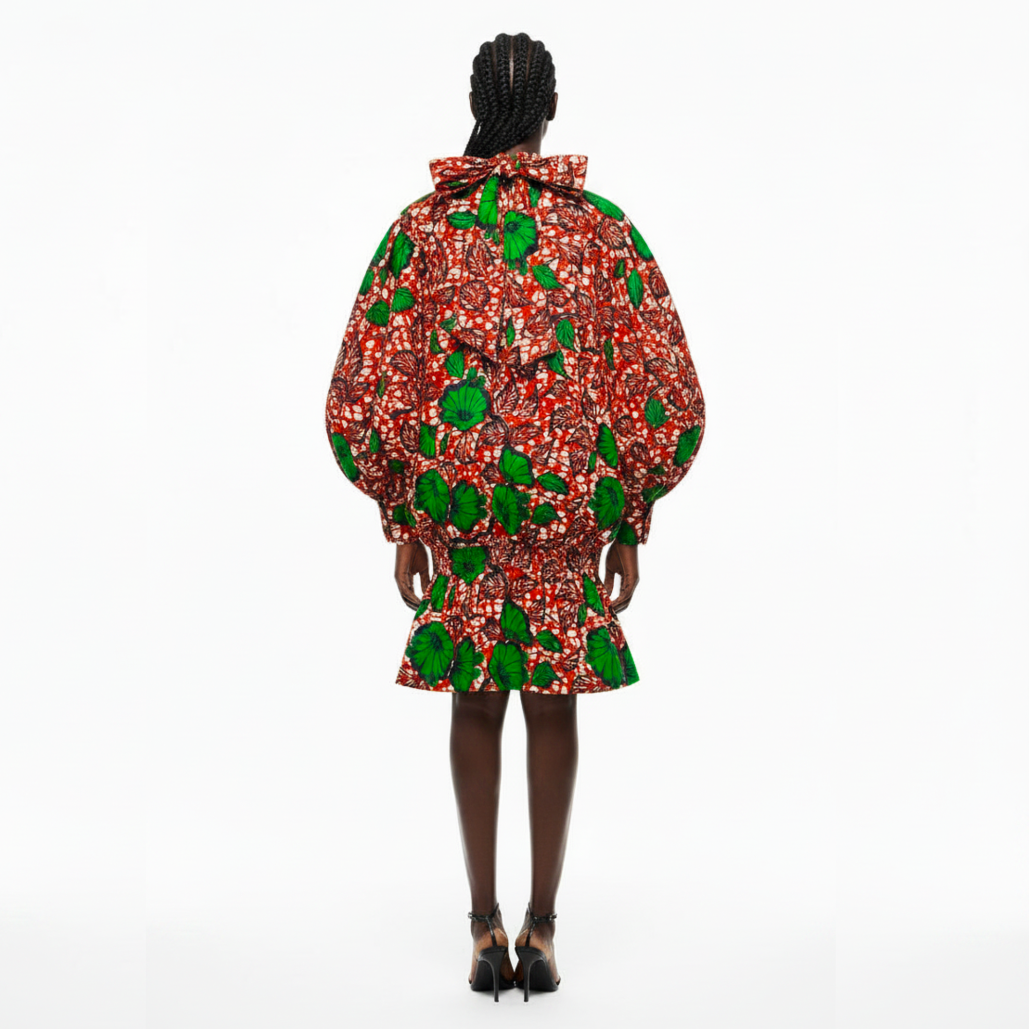 A model viewed from the back wearing a voluminous, short puff-sleeve dress with a bold red, green, and white abstract print, set against a plain white studio background.