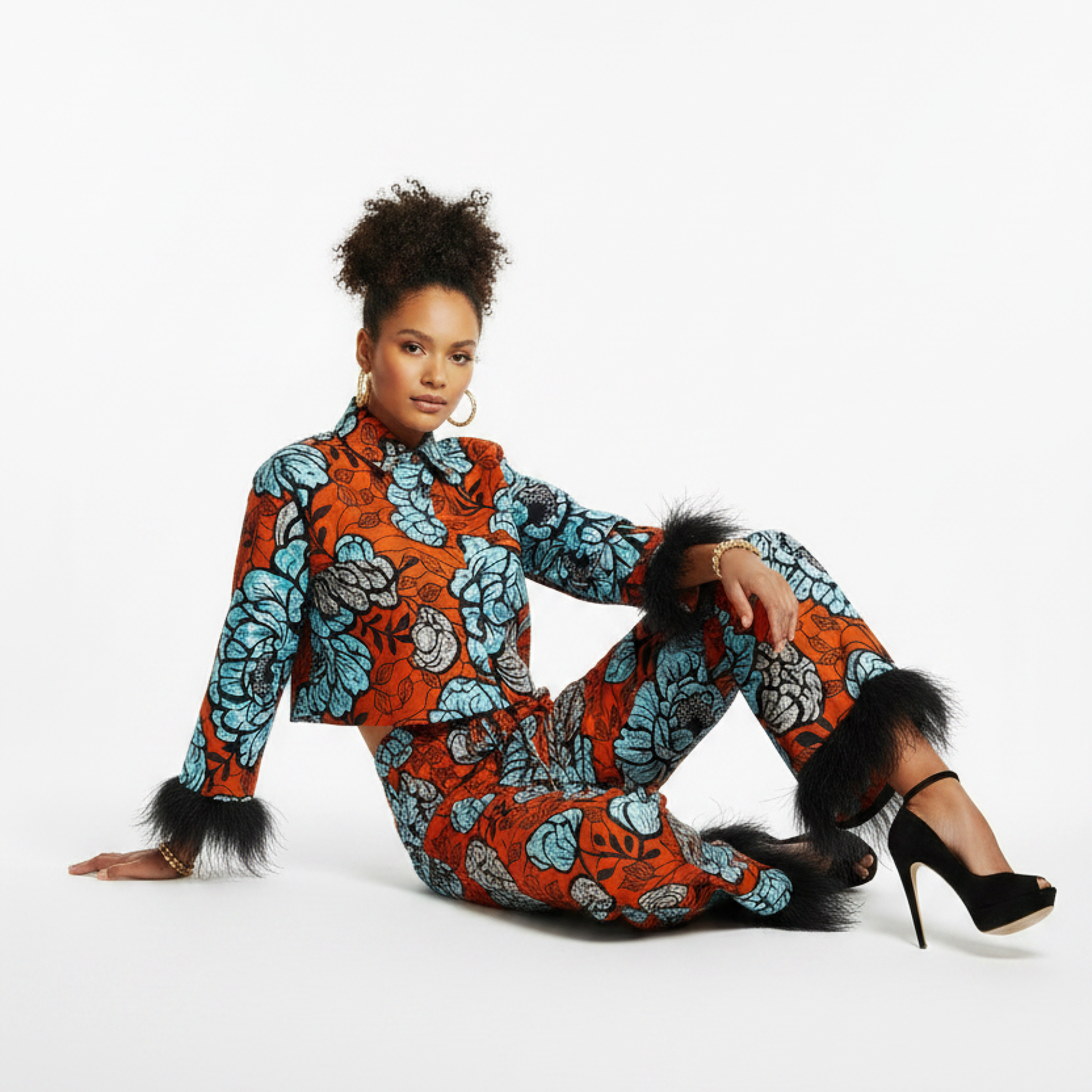 A model sits on a white studio floor wearing a matching orange and blue floral print two-piece set with black feather trim on the cuffs and hem, paired with black high heels.