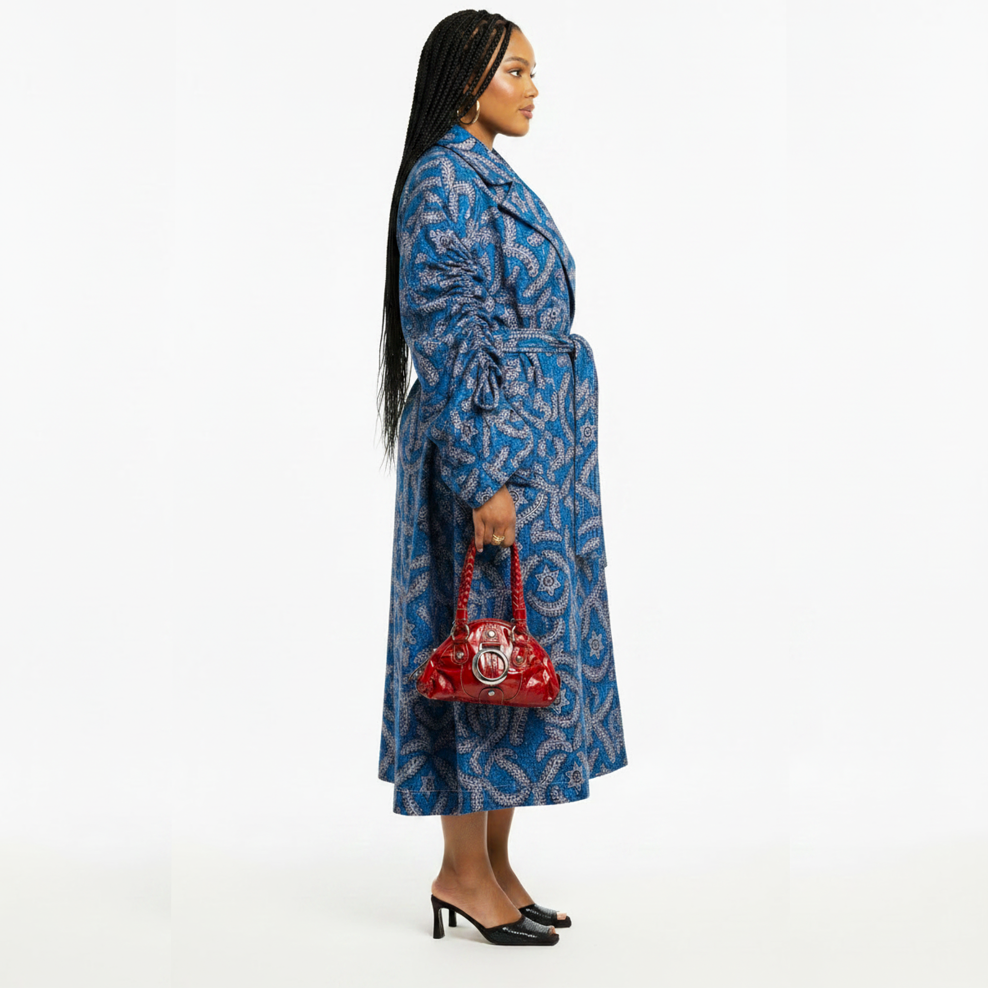 A woman stands in profile wearing a long blue patterned coat with a matching waist tie, holding a small red handbag, against a plain white studio background.