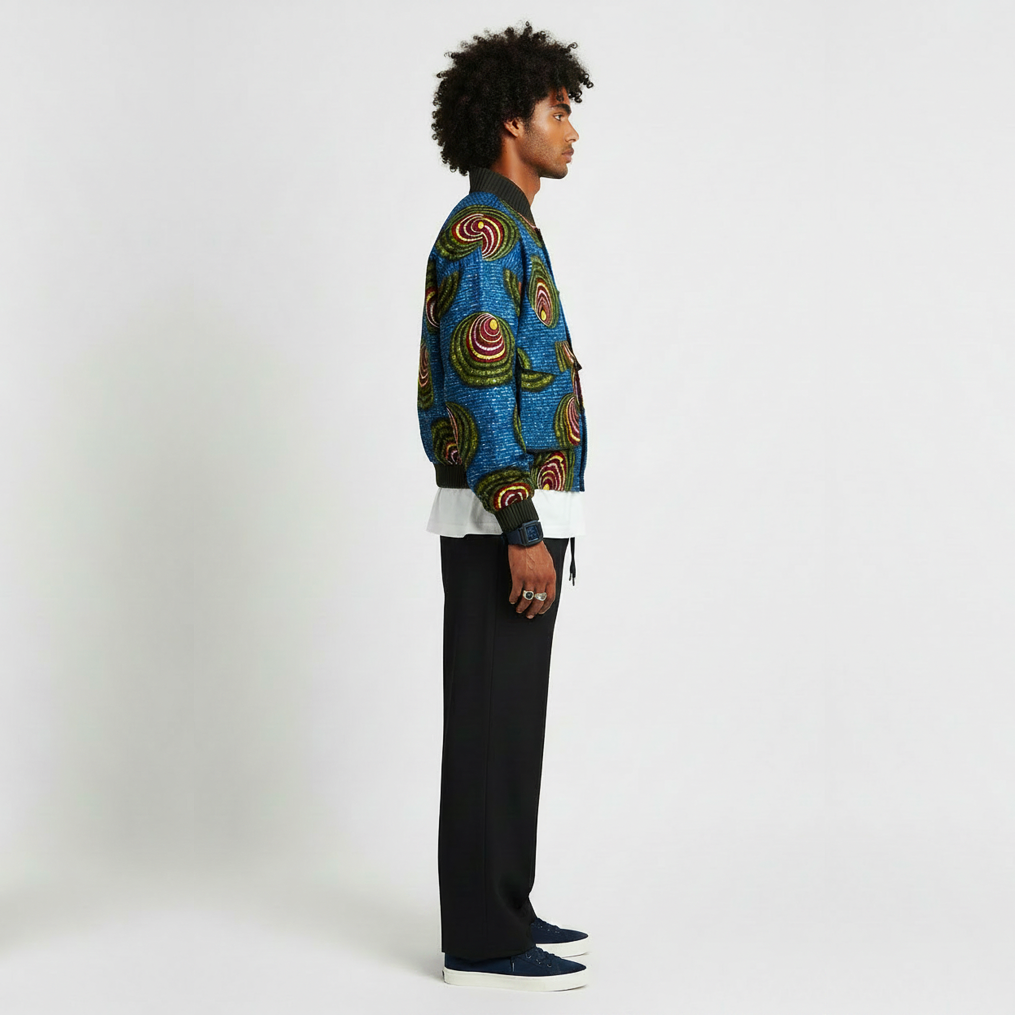 A man stands in profile wearing a blue patterned jacket with colorful spiral motifs, black trousers, and dark blue slip-on shoes against a plain light gray studio background.