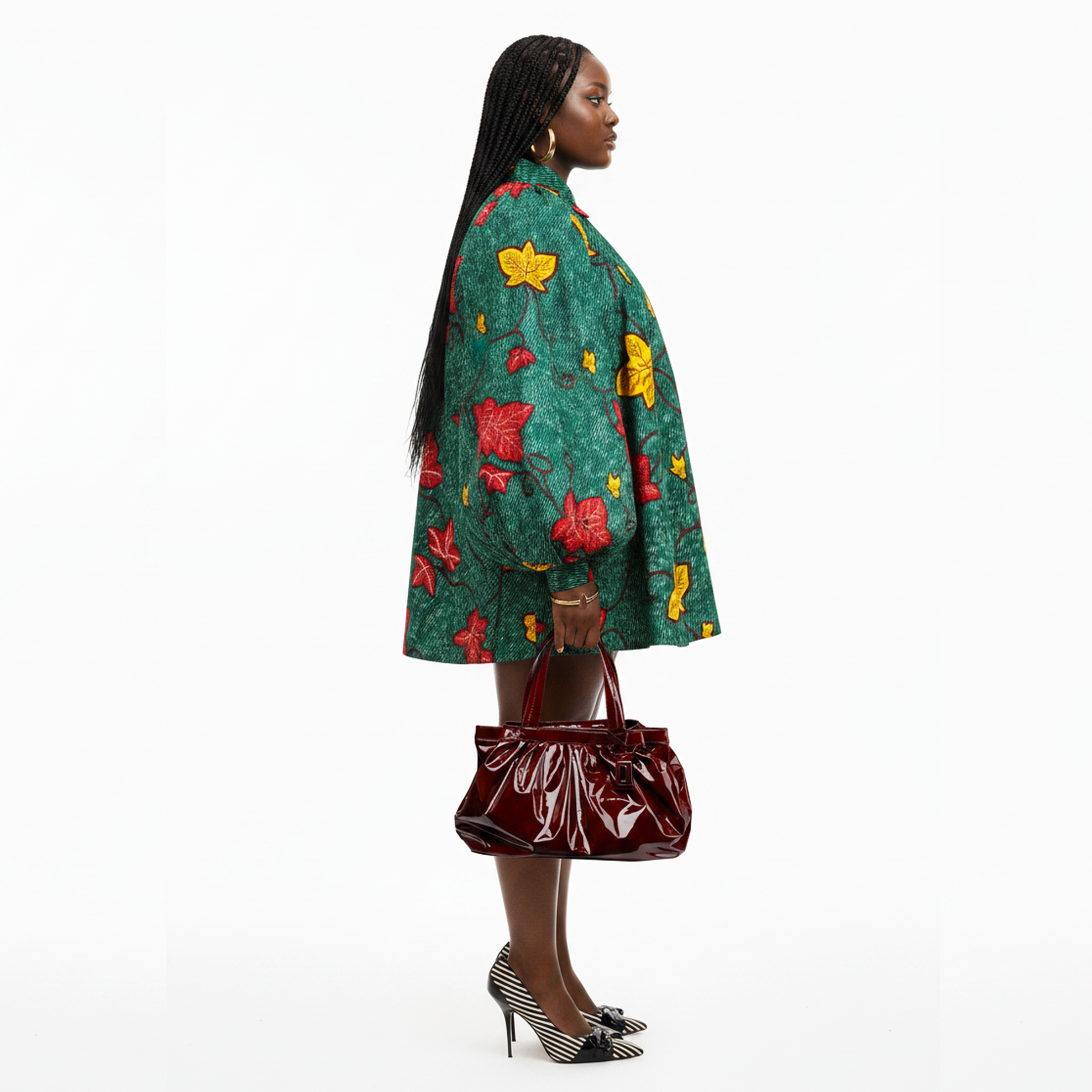 A woman stands in profile wearing a green patterned jacket with red and yellow leaf motifs, holding a shiny burgundy handbag, against a plain white studio background.