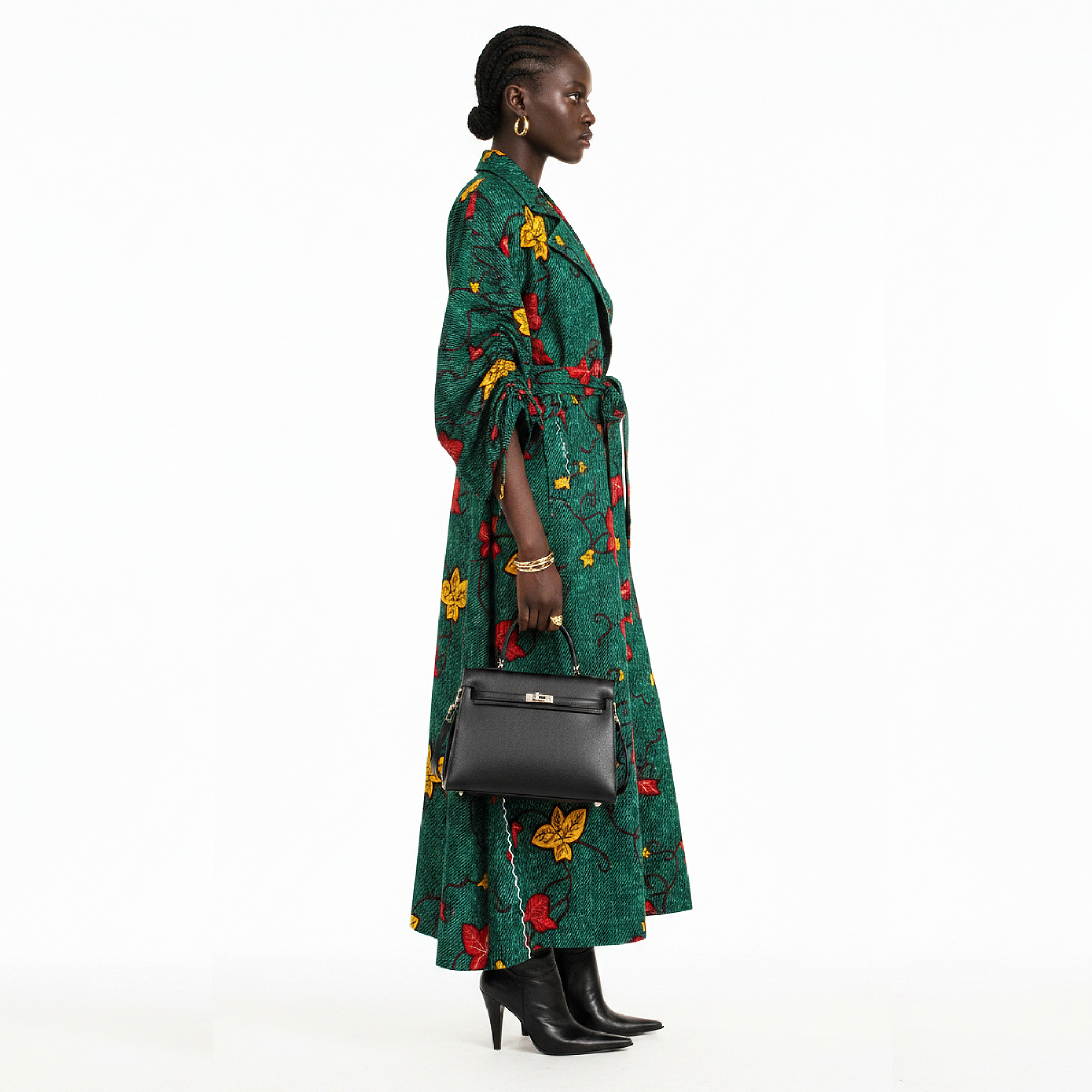 A woman stands in profile wearing a long green wrap-style coat with a bold red and yellow floral print, holding a black structured handbag, against a plain white studio background.