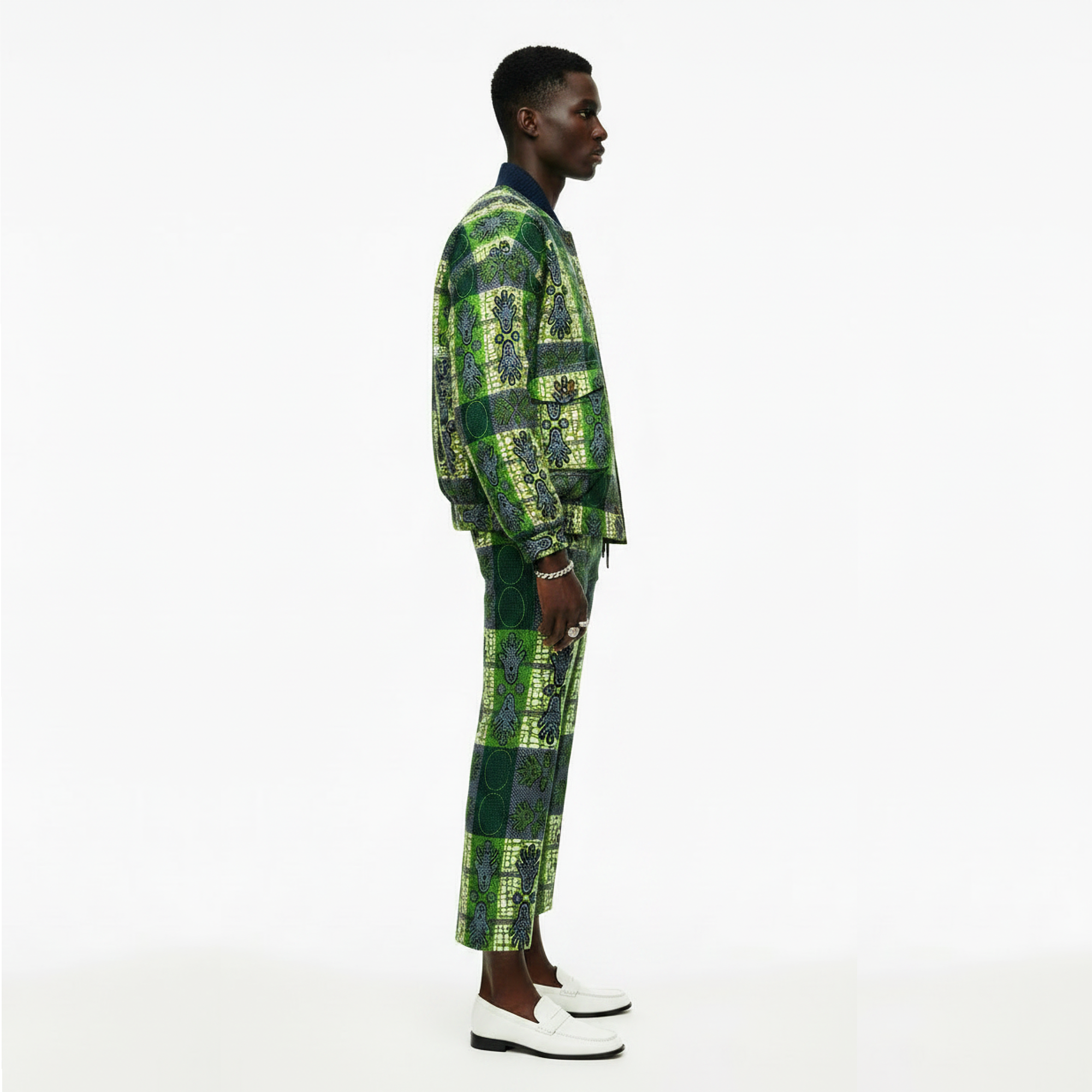 A man stands in profile wearing a matching green and black patterned two-piece outfit with a collared shirt underneath and white loafers, photographed against a clean white studio background.