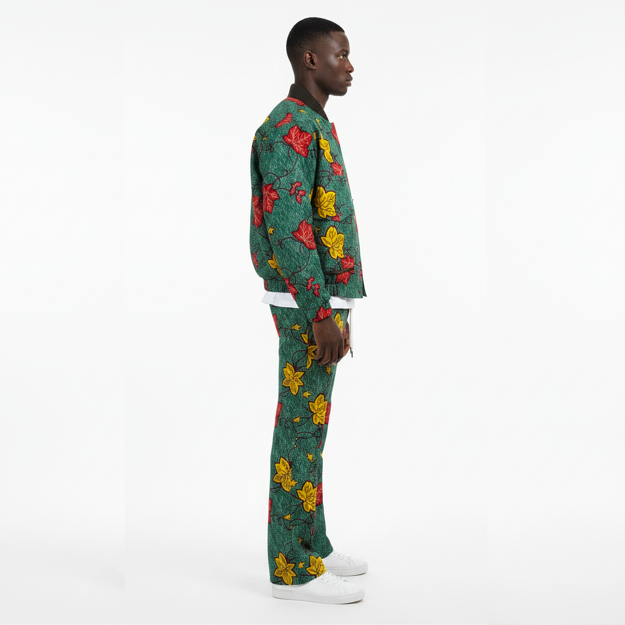 A man stands in profile wearing a matching green two-piece set with a red and yellow floral print, paired with white sneakers, against a plain white studio background.