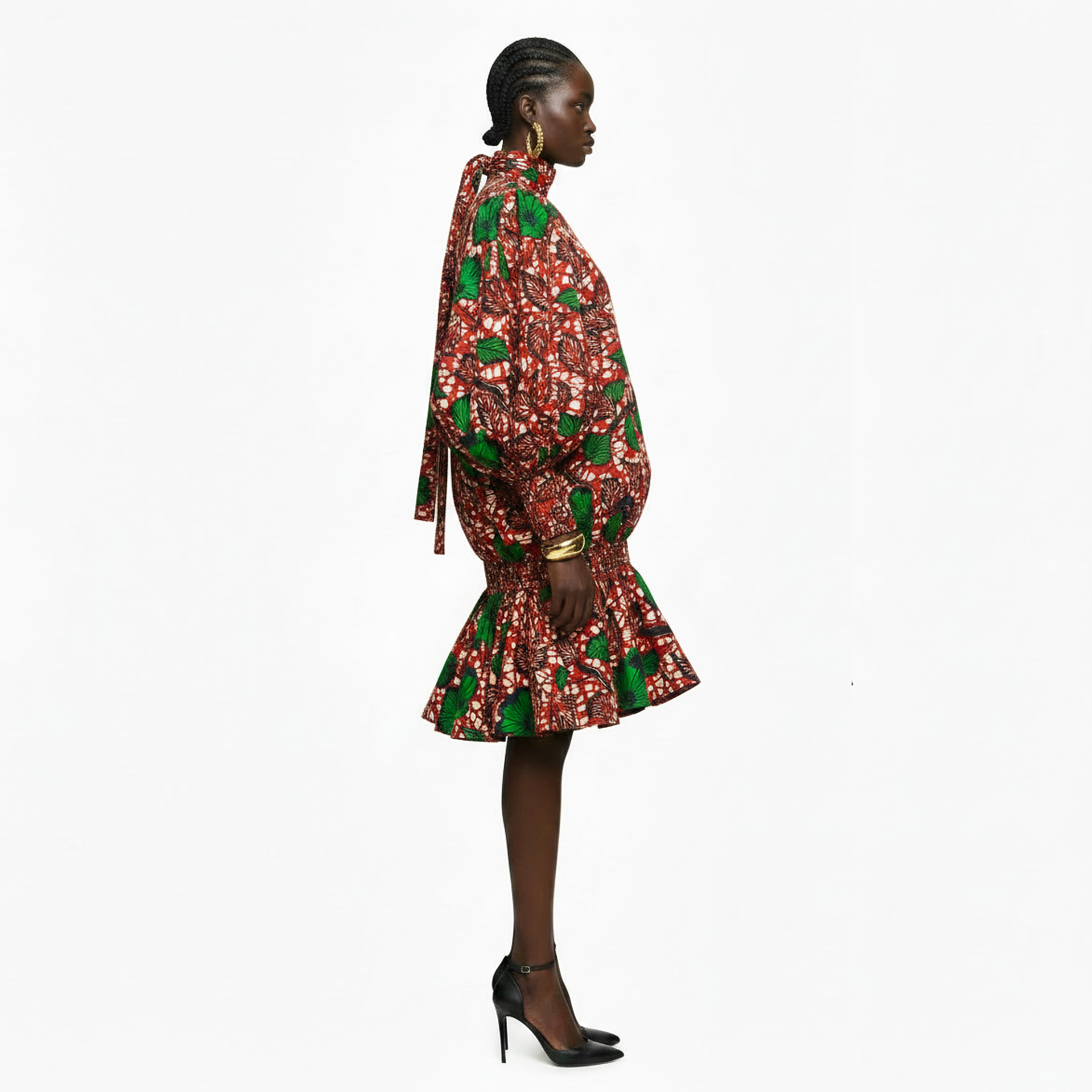A model is shown from the side profile wearing a short, voluminous mini dress with a red and green floral print, long balloon sleeves, and a tie-neck detail, paired with black high heels against a plain white background.