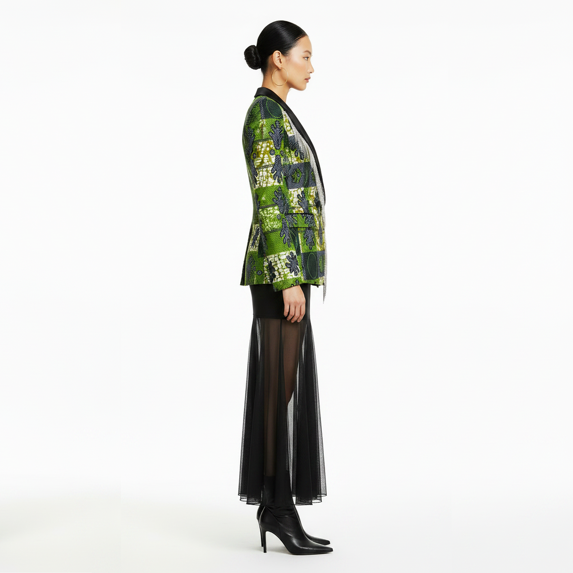 A model is shown in profile wearing a green and black patterned blazer, a sheer black midi skirt, and black high-heeled boots against a plain white studio background.