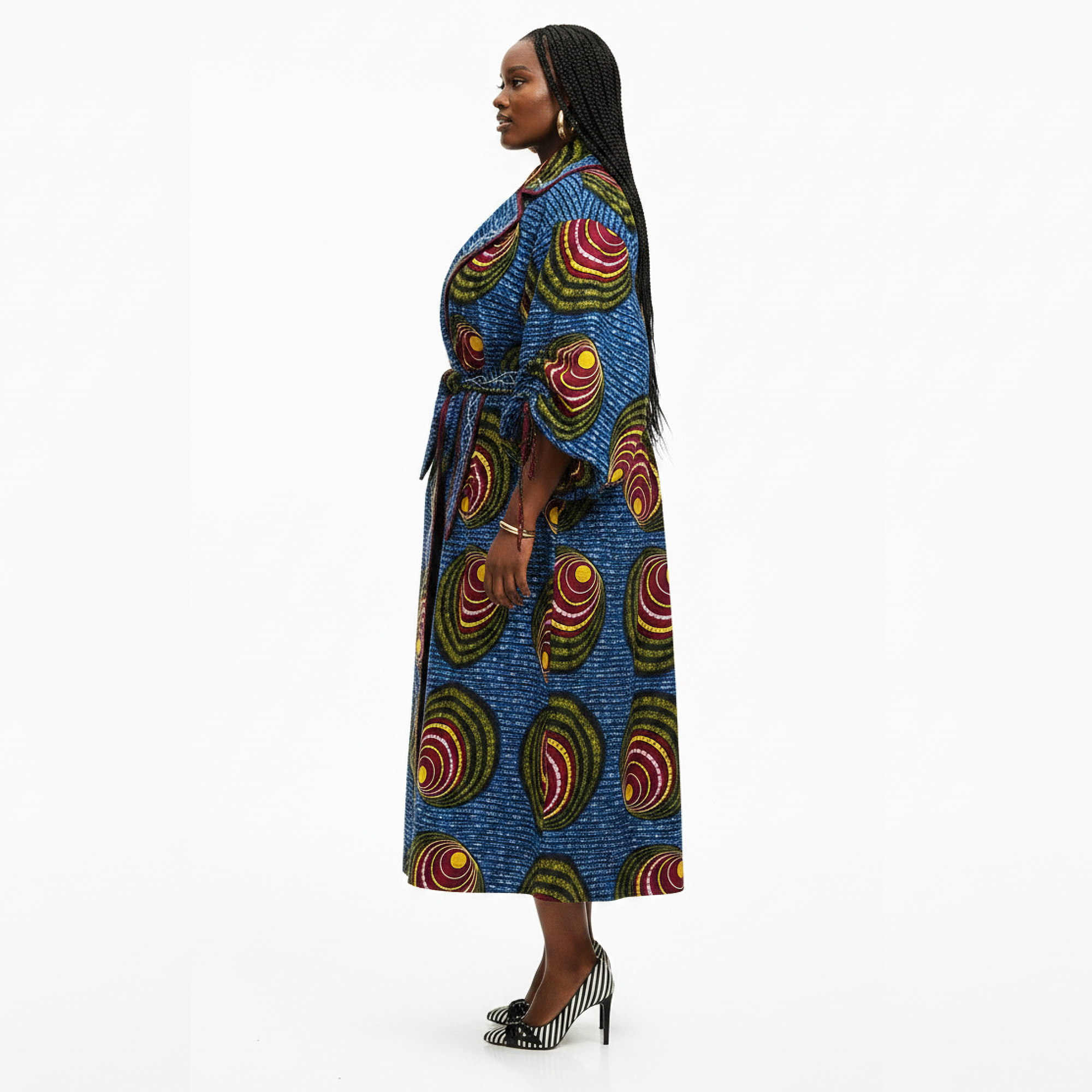 A woman modeling a long blue wrap coat with a vibrant African wax print pattern featuring red, yellow, and green concentric circles, shown in profile against a white studio background.