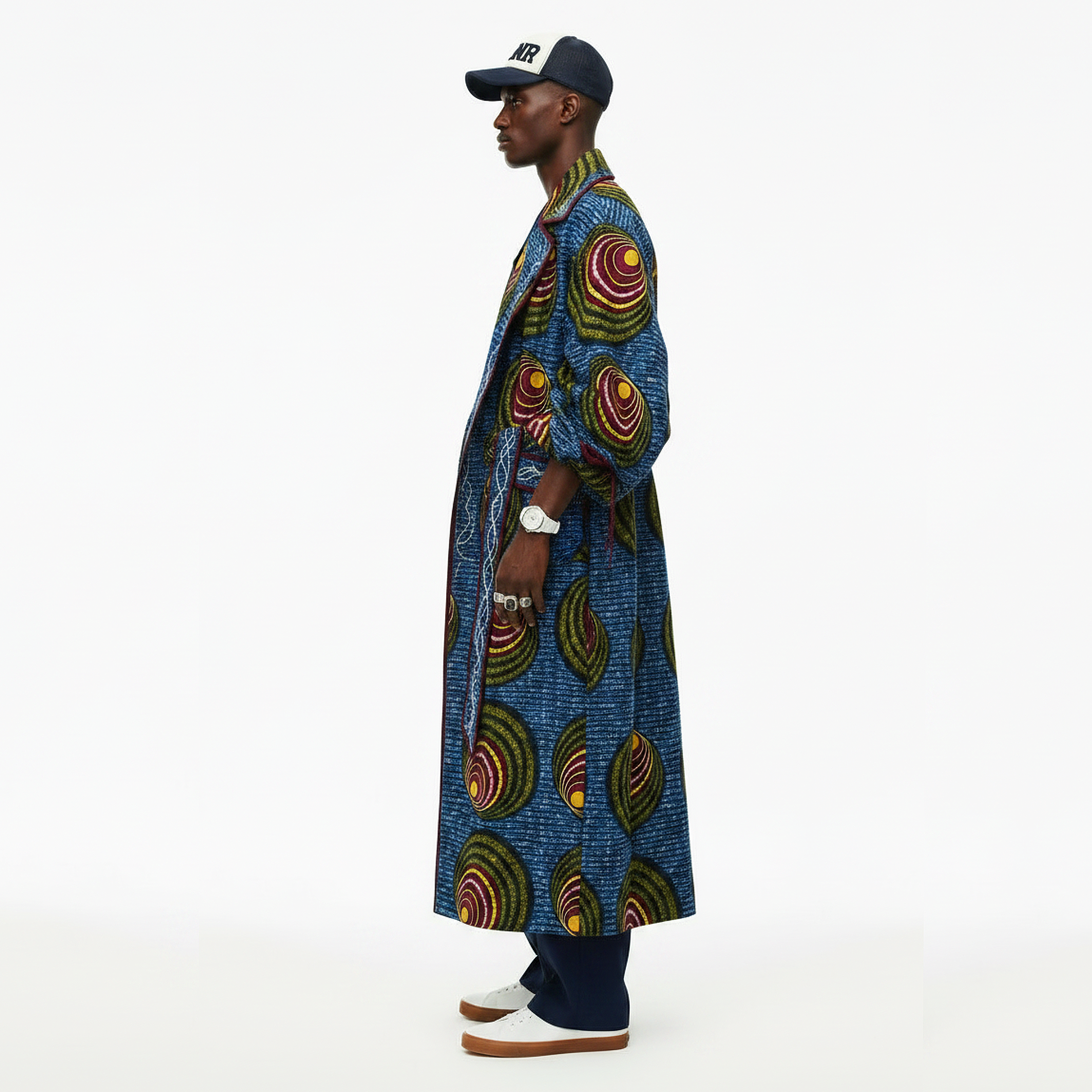A man in profile wearing a long blue patterned coat with circular multicolor motifs, navy pants, white shoes, and a navy cap, photographed against a clean white studio background.