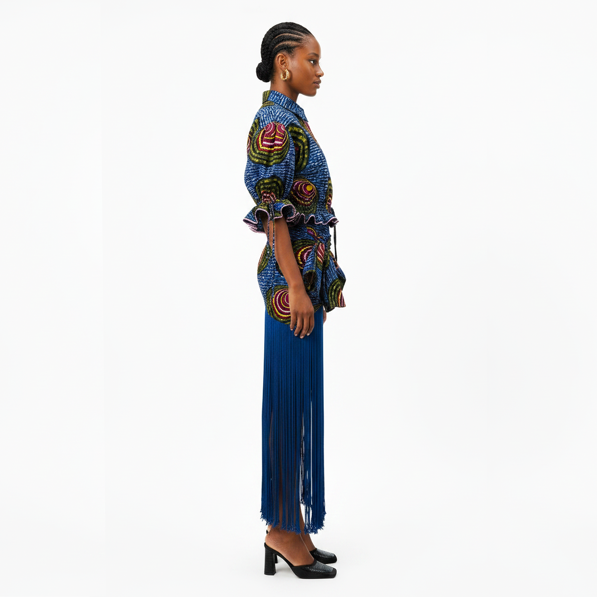 A woman shown in profile wearing a colorful patterned blouse and a long blue fringe skirt against a plain white studio background.