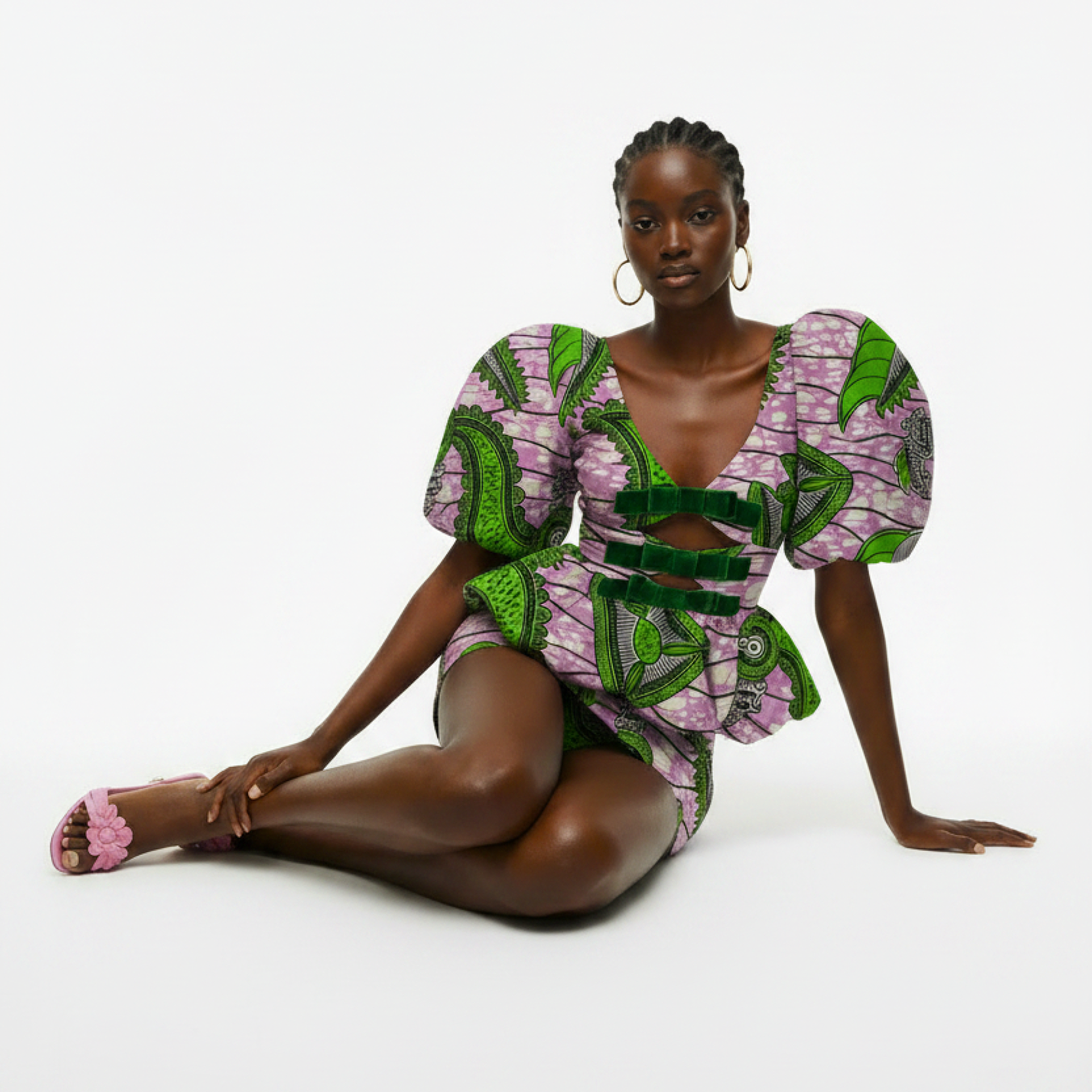 A model wearing a short puff-sleeve dress with a green and pink patterned African wax print, sitting on a white studio background.