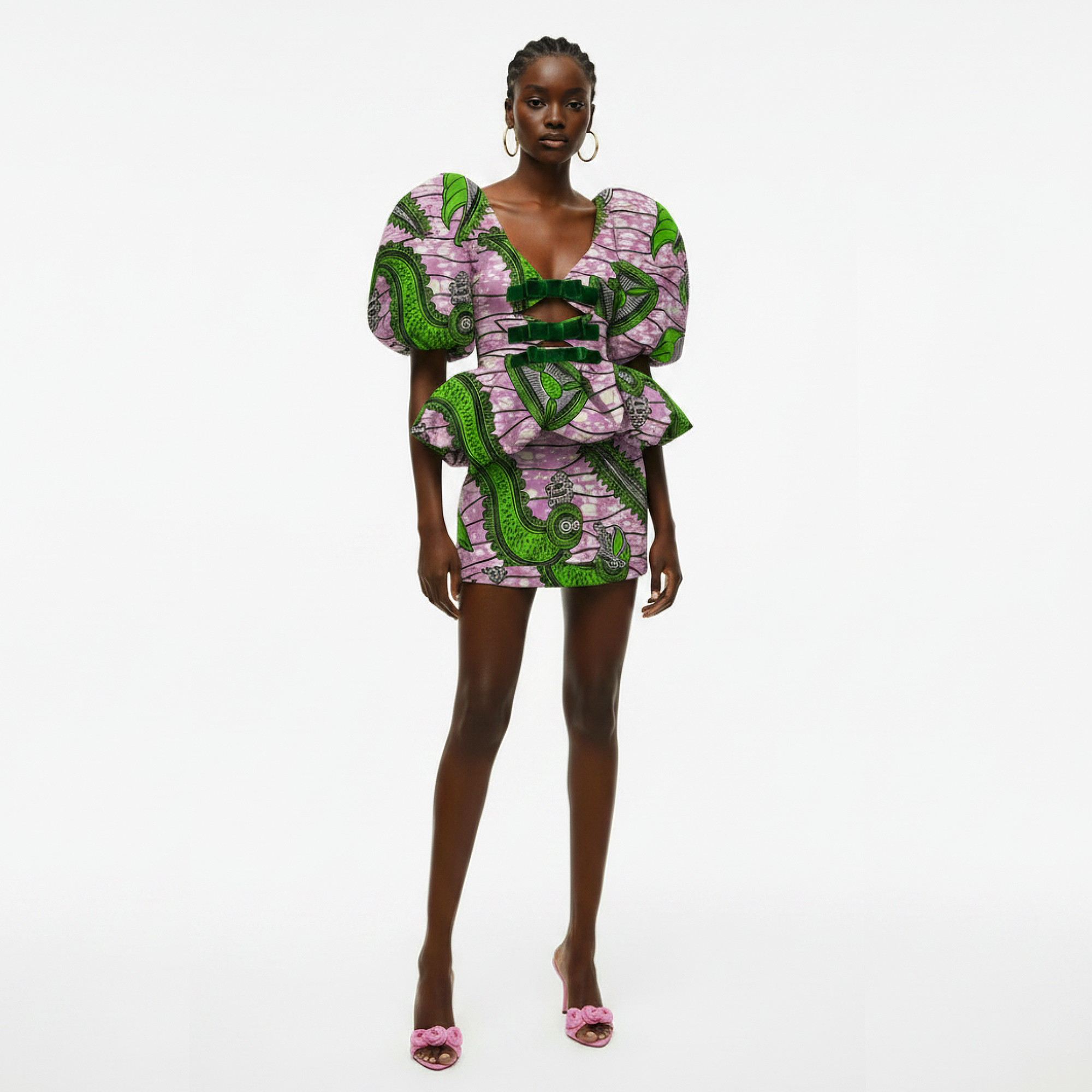 A model stands against a plain white studio background wearing a short puff-sleeve dress with a bold green and pink patterned print and a front cutout. She is also wearing pink heeled sandals and large hoop earrings.