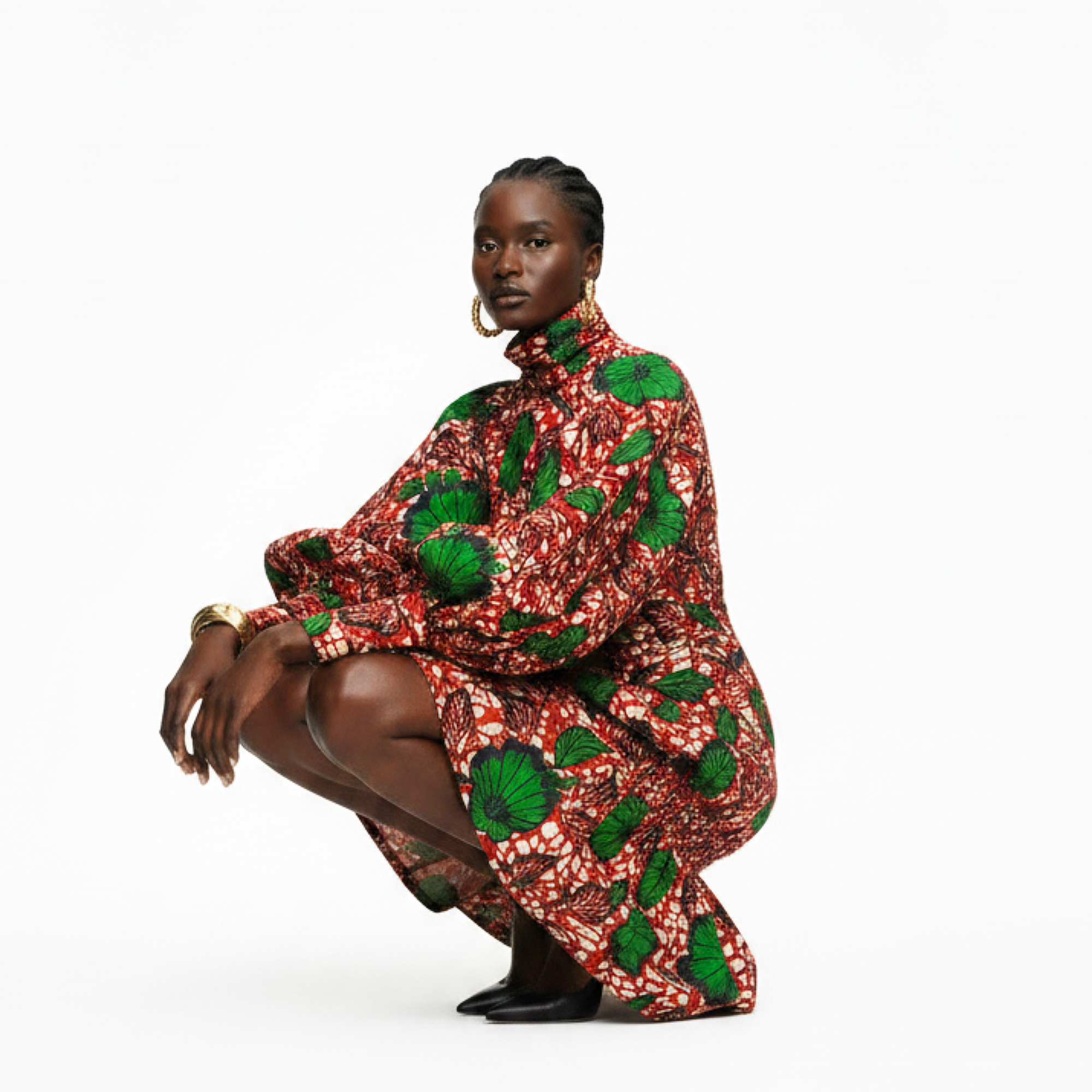 A model crouches in a high-neck, voluminous dress with a bold red, white, and green leaf pattern, set against a solid white studio background.
