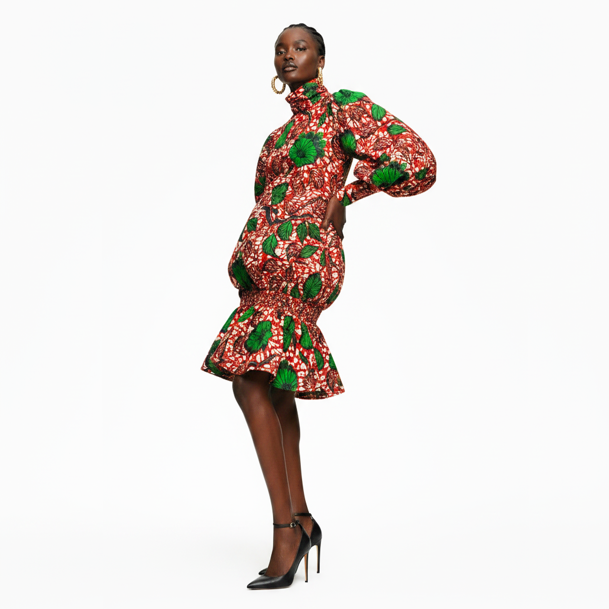 A model wearing a high-neck, puff-sleeve mini dress with a red, white, and green floral print, paired with black high heels, against a solid white background.