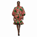 A model wearing a high-neck, long-sleeved mini dress with a vibrant red, green, and white floral print, set against a solid white background.