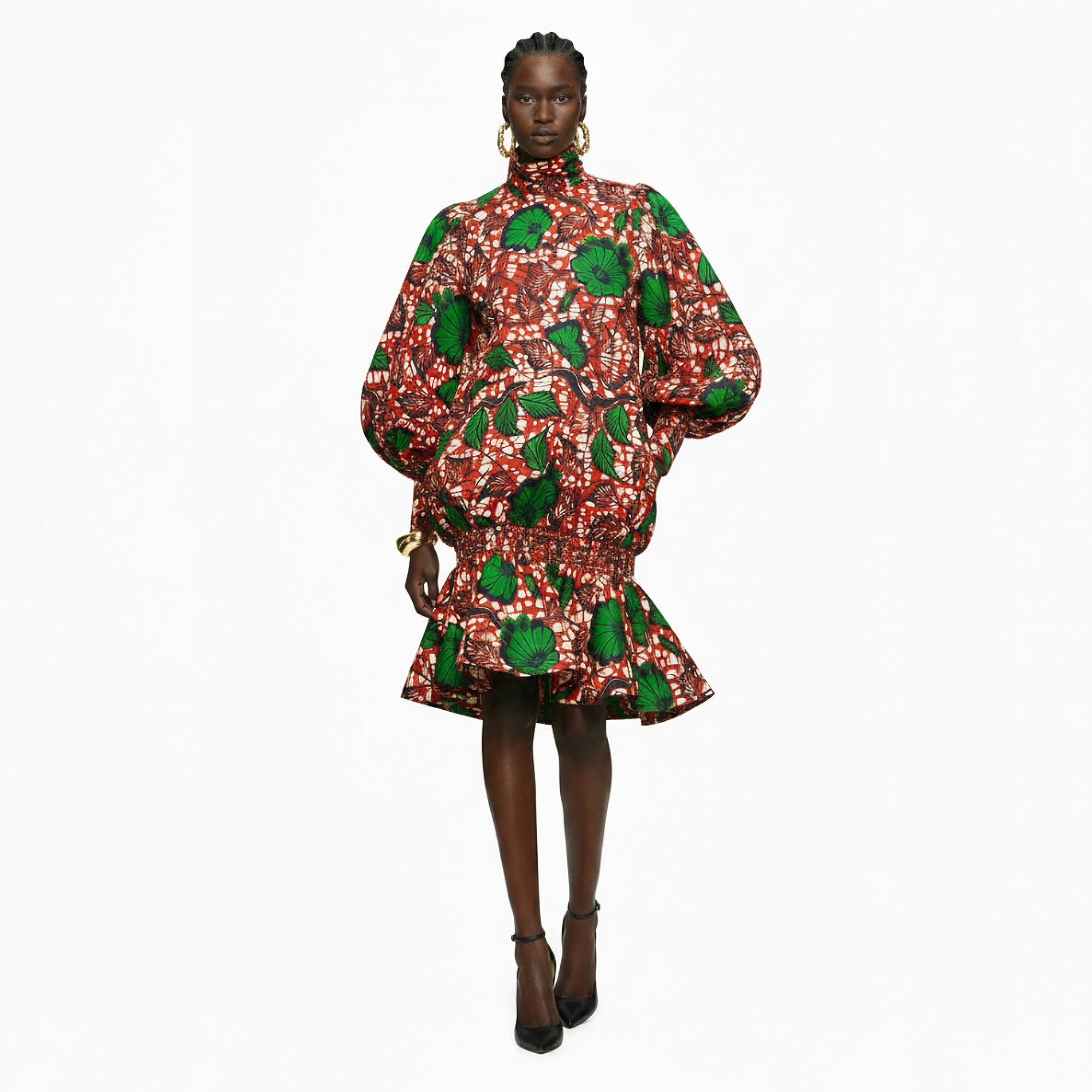 A model wearing a high-neck, long-sleeved mini dress with a bold red, white, and green floral print, styled with gold hoop earrings and black heels against a plain white background.