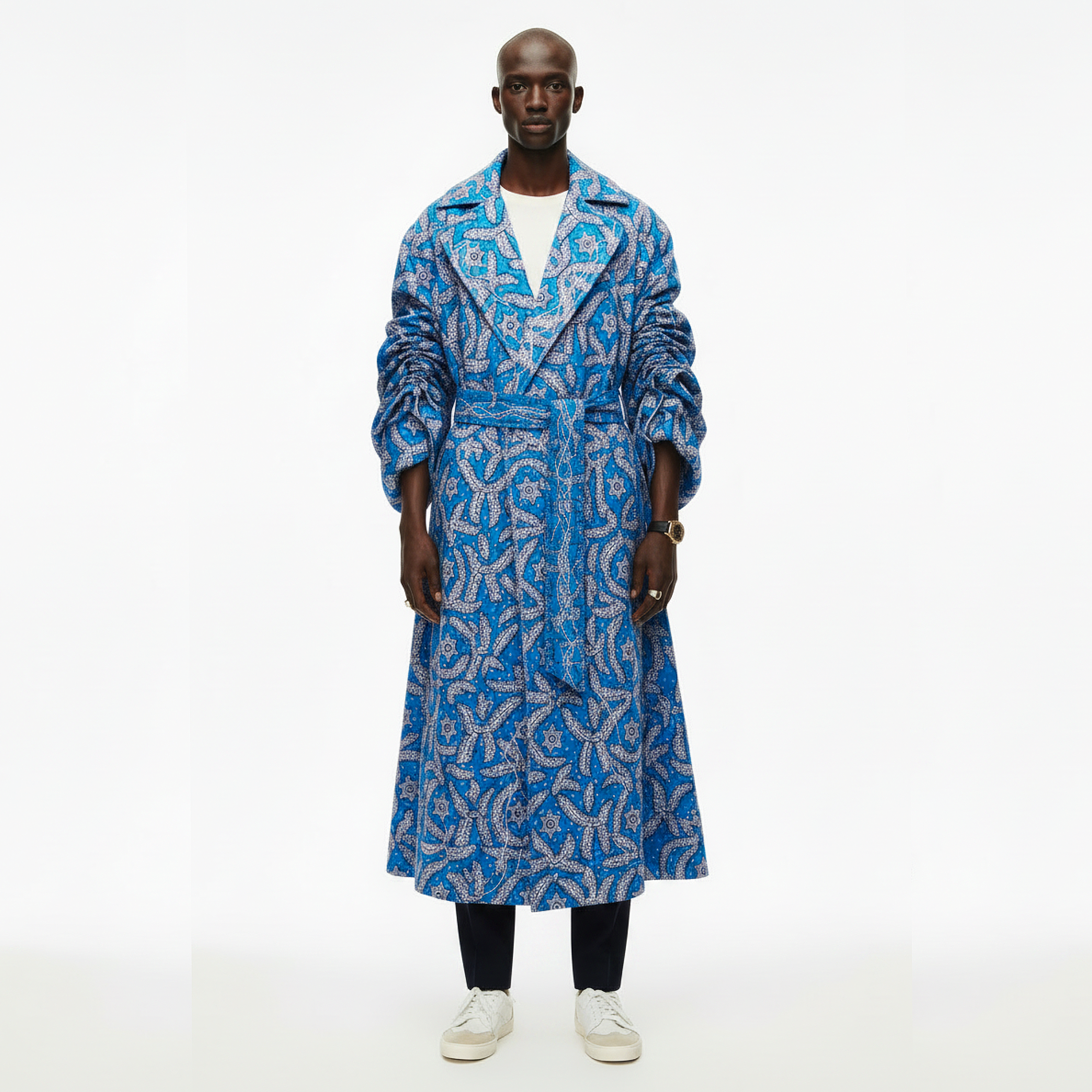 A model stands against a plain white studio background wearing a long blue coat with a bold white geometric pattern, a white t-shirt, black pants, and white sneakers.