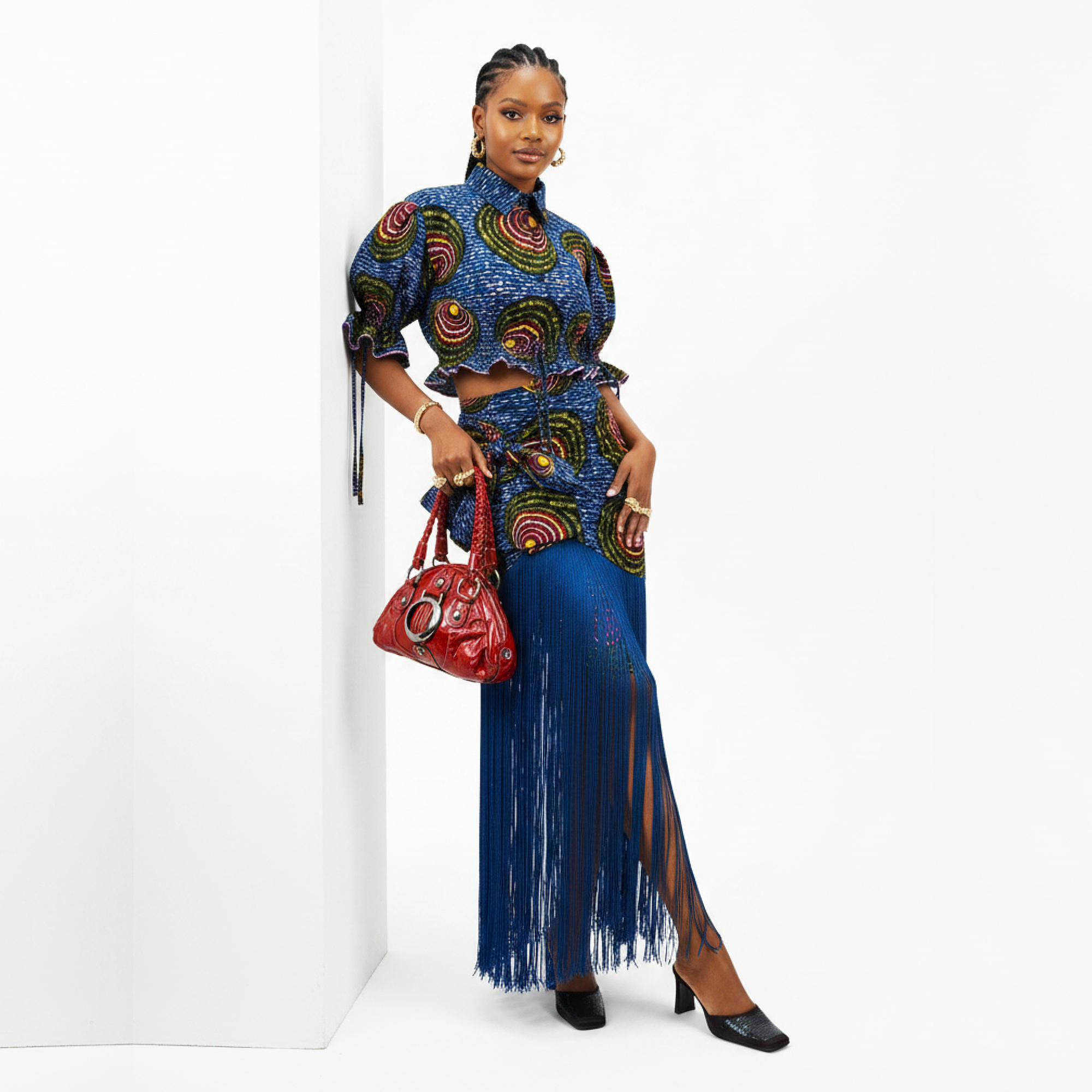 A model stands in a white studio corner wearing a blue patterned two-piece set with a high-neck long-sleeve top and a matching long skirt featuring fringe. She is holding a red handbag and wearing black heels.
