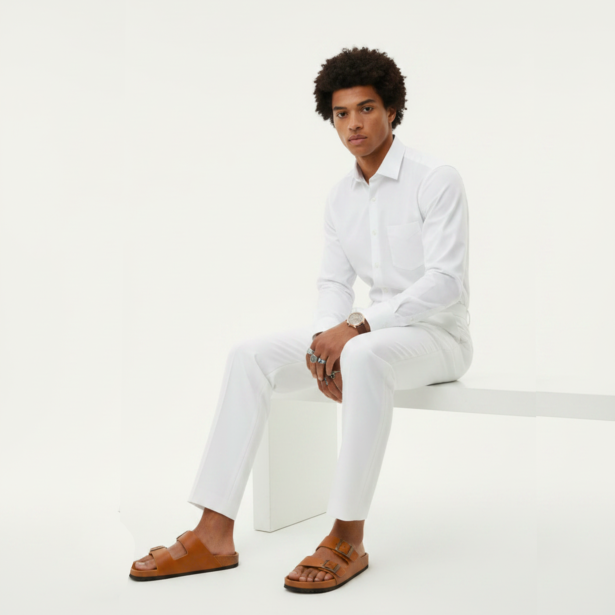 Man wearing a white shirt and pants with brown sandals sitting on a white surface.
