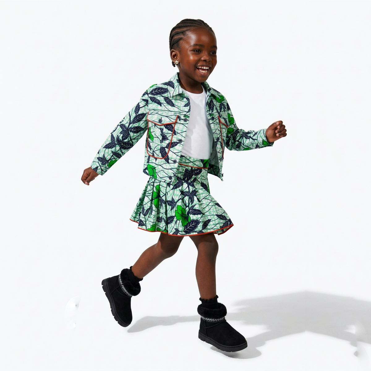A young girl is shown in motion, wearing a matching green and black leaf-patterned jacket and skirt set with a white t-shirt underneath and black boots. She is posing on a clean white studio background.