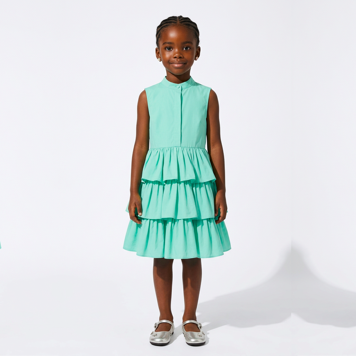 A young girl with braided hair stands against a white studio background wearing a sleeveless tiered dress in a bright mint green color, paired with metallic silver shoes.