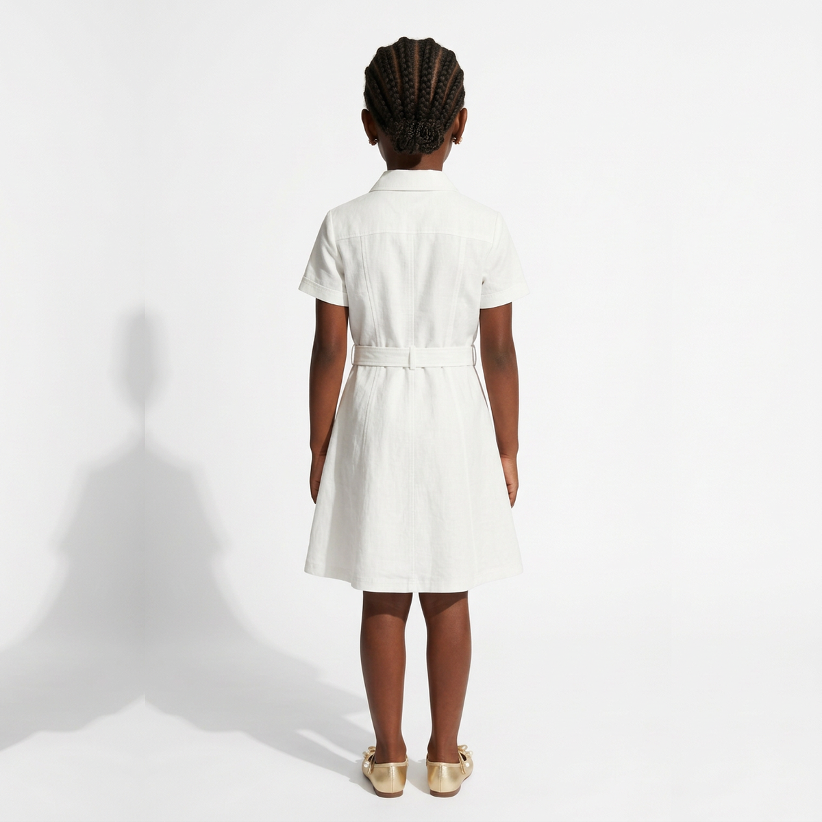 A young girl viewed from behind wearing a white short-sleeve collared dress with a matching belt and metallic gold shoes, standing against a white studio background.
