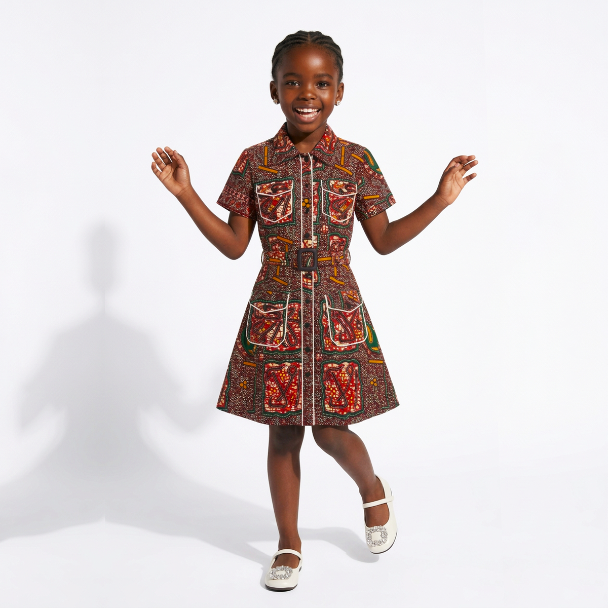 A young girl smiling and posing in a short-sleeve collared dress with a colorful, busy African wax print pattern featuring red, brown, green, and yellow motifs, set against a plain white studio background.