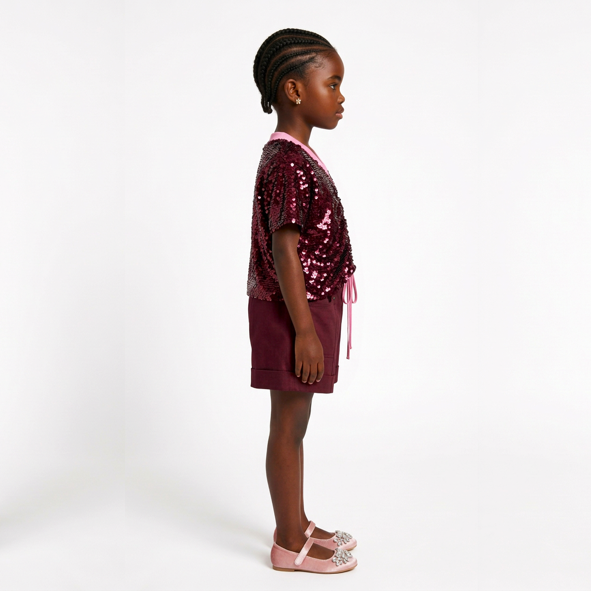 A young girl standing in profile wearing a short-sleeve sequined top and matching shorts in a deep burgundy shade, paired with light pink flat shoes, against a plain white studio background.