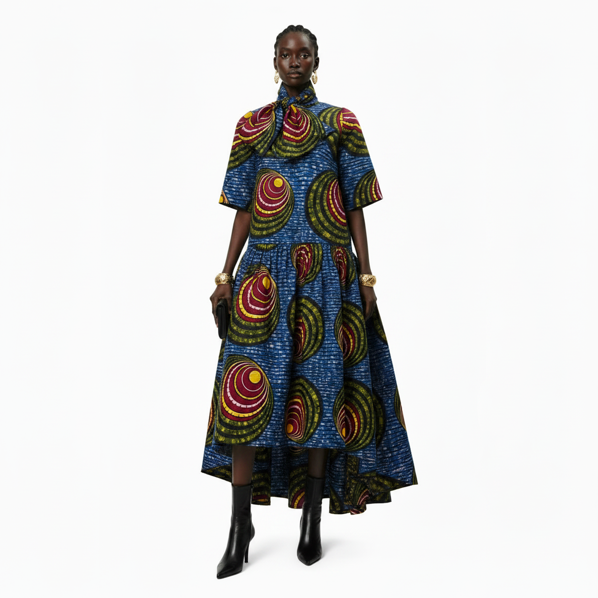 A model stands against a plain white studio background wearing a short-sleeve high-low midi dress with a vibrant blue base and a bold multicolor circular pattern in red, yellow, green, and black. She is also wearing black ankle boots and gold jewelry.