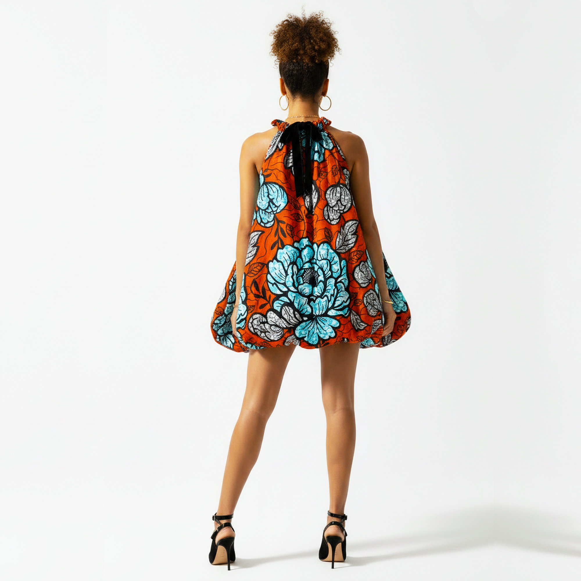 A woman viewed from behind wearing a sleeveless halter mini dress with a vibrant orange, blue, and gray floral pattern, paired with black high-heeled sandals against a plain white studio background.