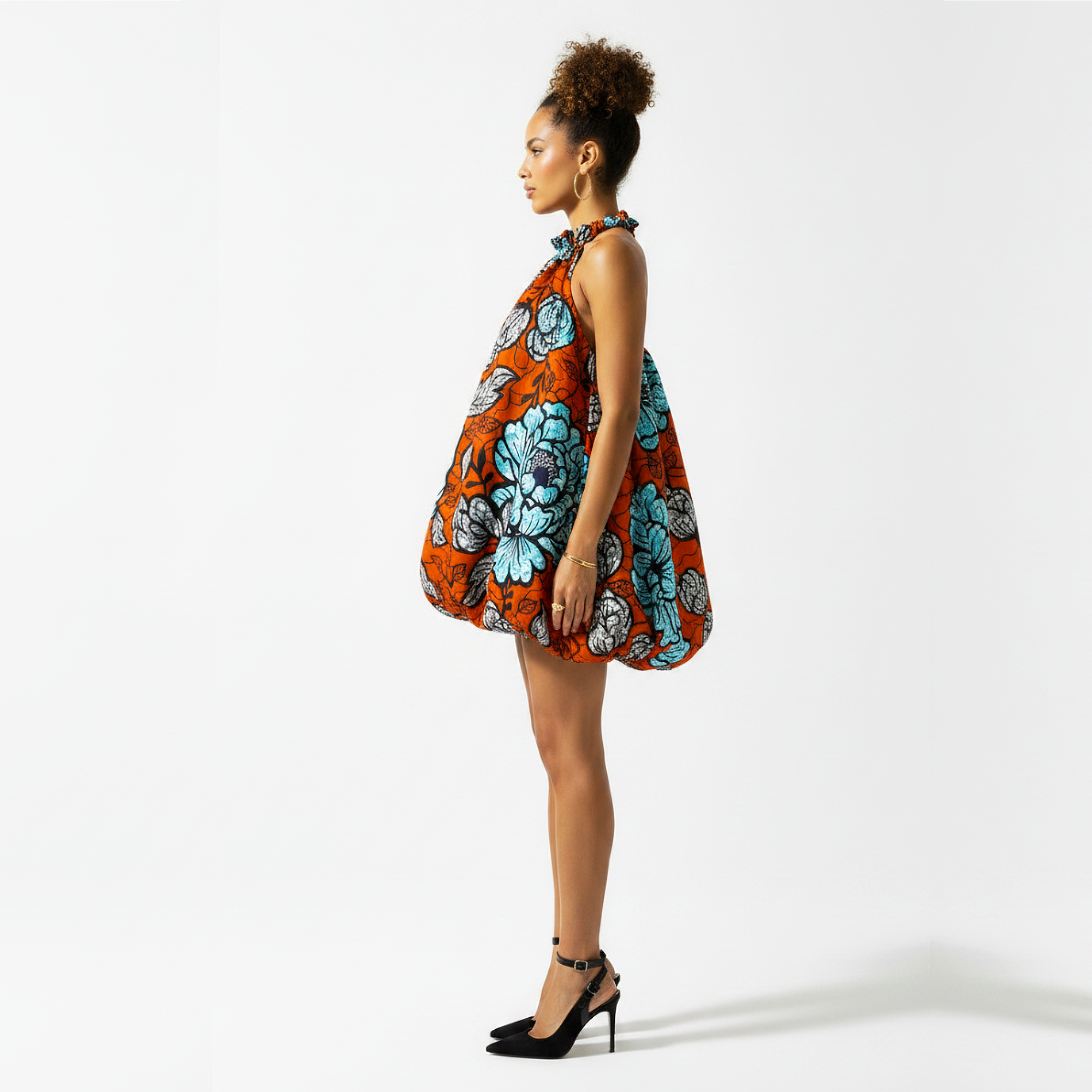 A woman stands in profile wearing a sleeveless, balloon-style dress with a bold orange, turquoise, and gray floral print, paired with black high heels, against a plain white studio background.