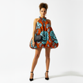 A model wearing a vibrant orange halter mini dress with a bold blue and gray floral print, holding a black quilted handbag, set against a plain white studio background.