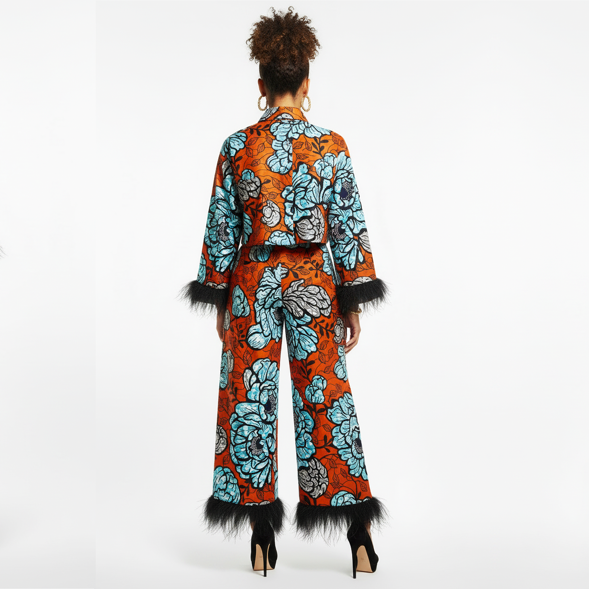 A model viewed from behind wearing a matching two-piece set consisting of a cropped long-sleeve top and wide-leg pants, both featuring a vibrant orange and light blue floral print with black feather trim at the cuffs and hems.