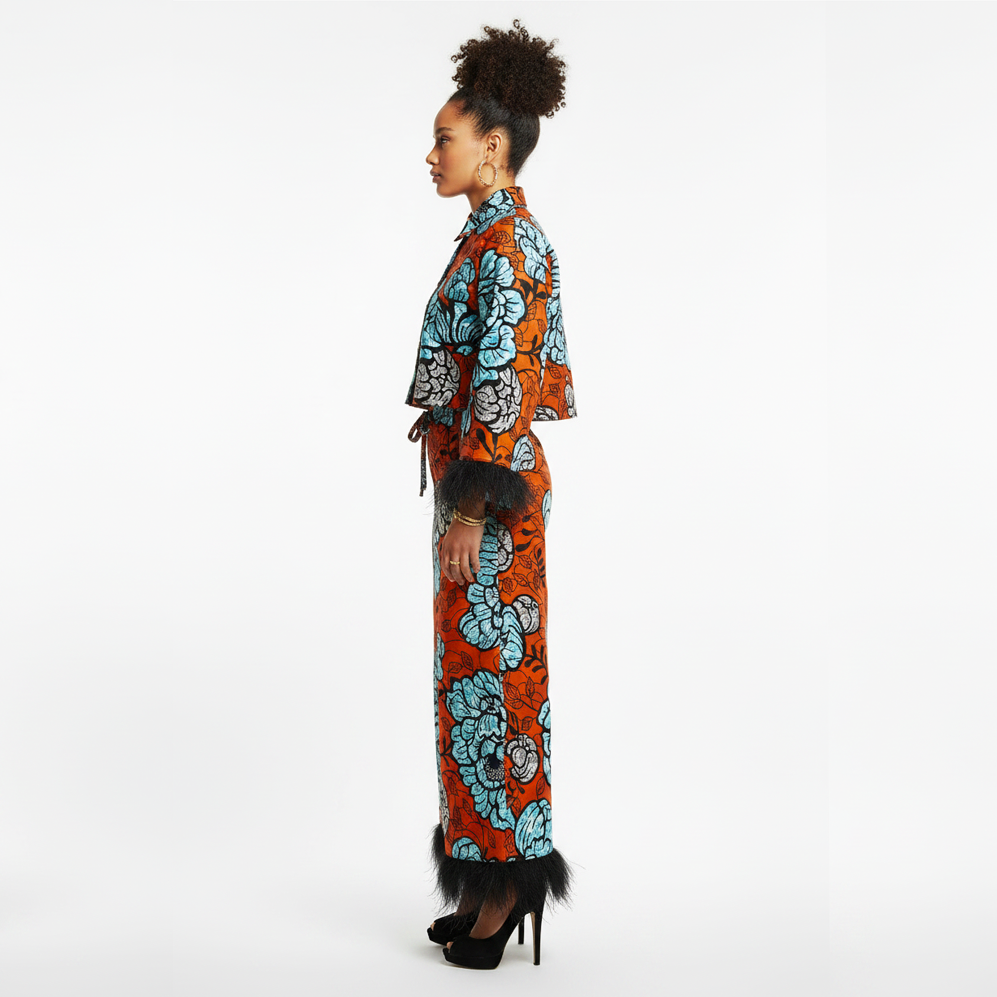 A woman stands in profile wearing a long orange dress with a bold blue and black floral pattern, black feather trim at the cuffs and hem, and black high heels against a plain white studio background.
